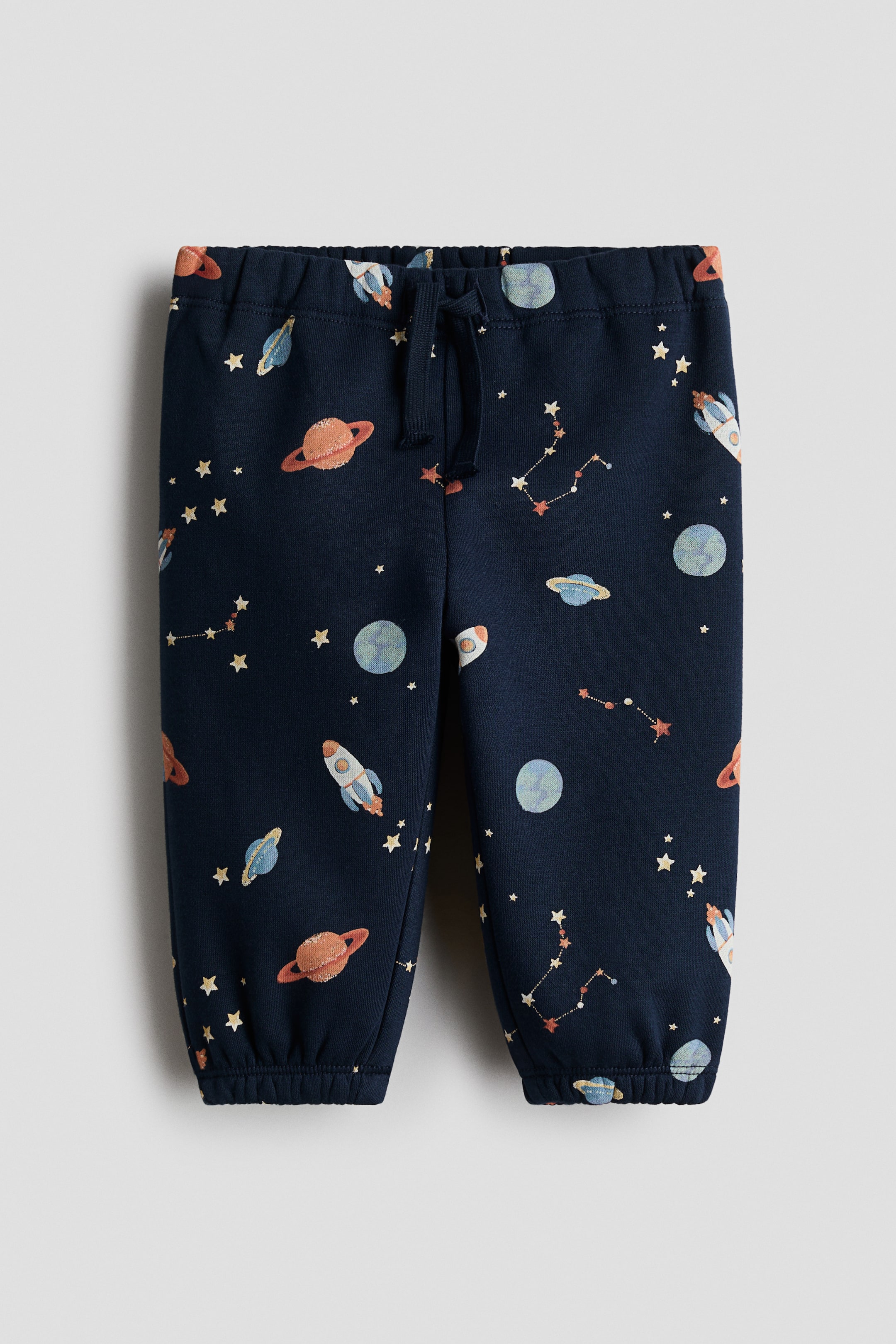 View larger image: Navy blue joggers crafted from sweatshirt fabric showcase a space-themed print with planets, rockets, and stars. Designed with an elastic waist that includes a decorative drawstring, they feature gathered hems with covered elastic.