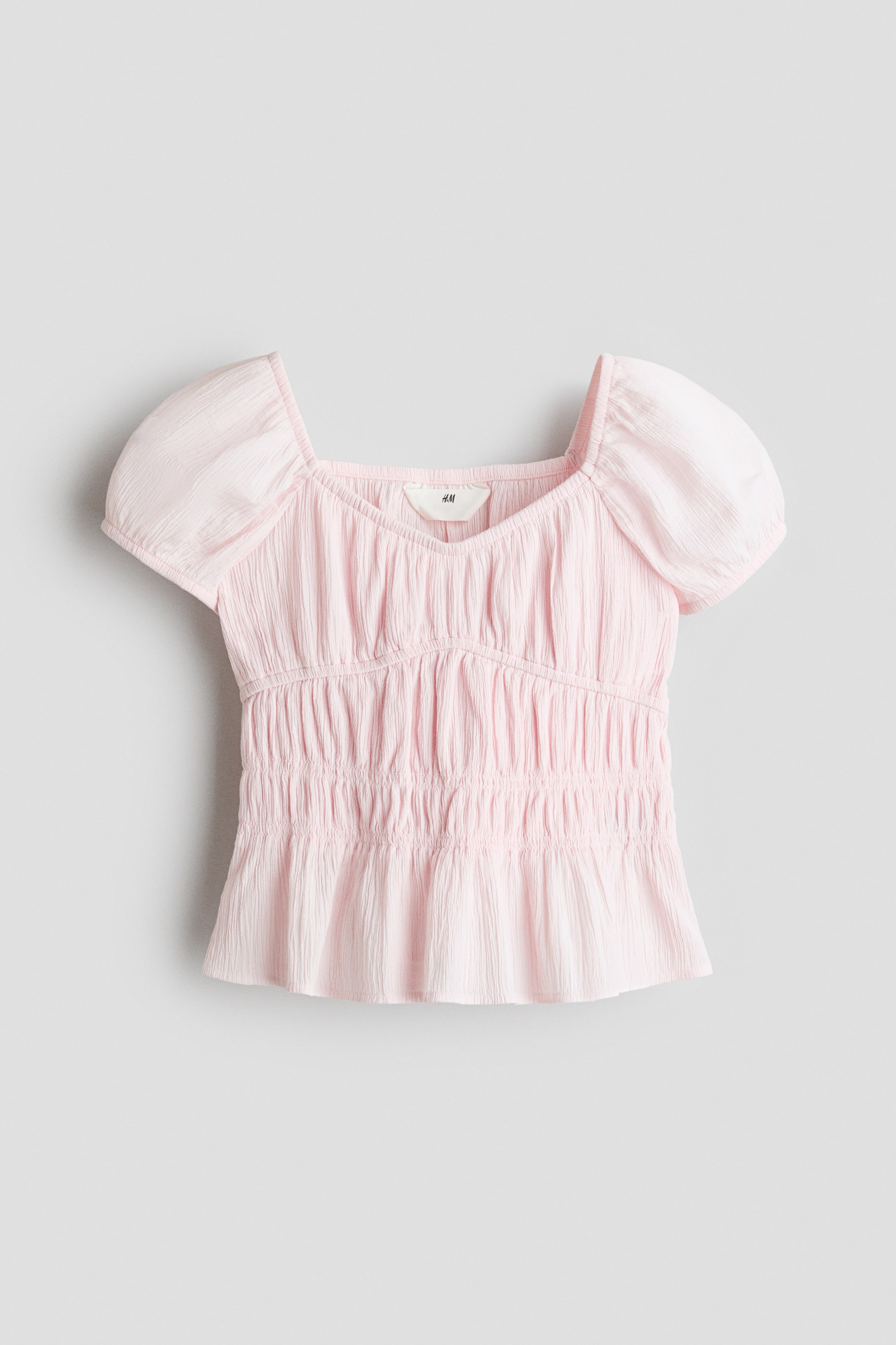 View larger image: A light pink top is showcasing its front view, featuring a square neckline with a subtle V-shape at the center, and short puff sleeves. The crinkled fabric highlights smocking detail across the bodice, creating a fitted waist and a slightly flared hem.