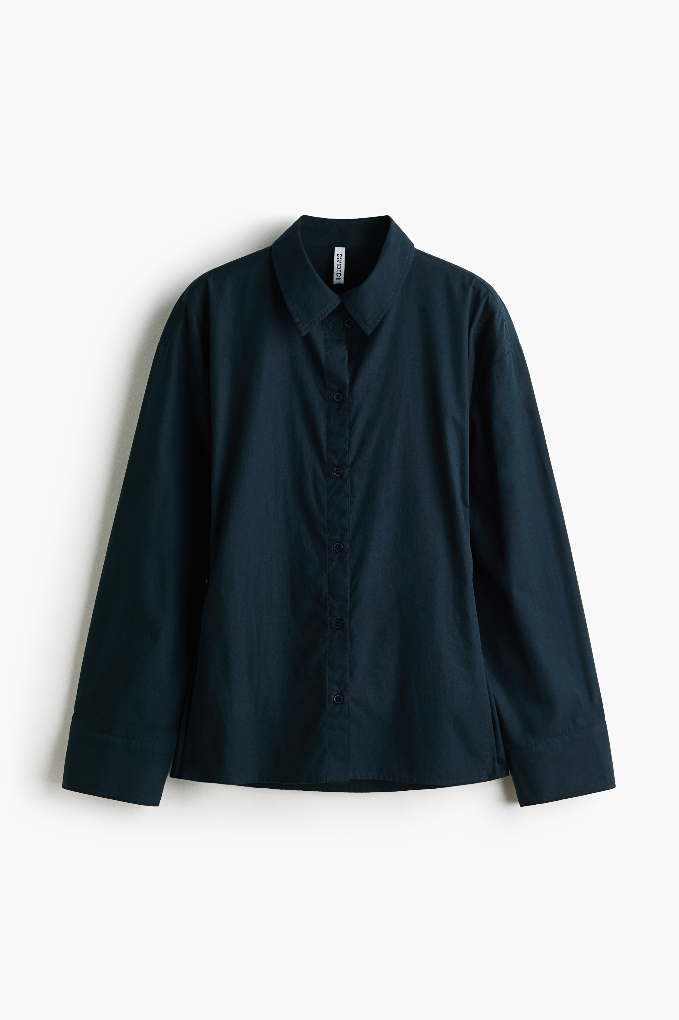 Una camicia in popeline di cotone blu navy scuro è mostrata di profilo laterale rivolto a sinistra, con colletto classico, spalle scese e maniche lunghe con polsini con bottoni. La camicia è indossata abbottonata, con una silhouette morbida che cade su pantaloni a gamba larga.