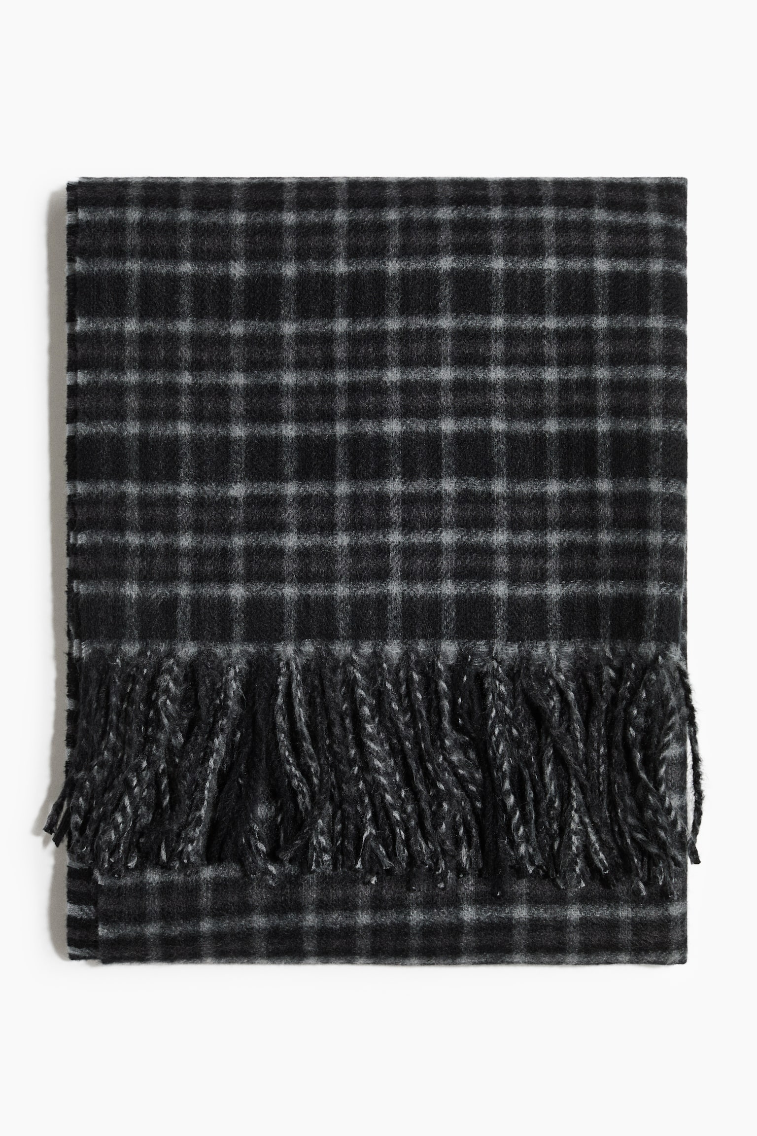 A folded scarf, shown from the front, displays a black and grey tartan pattern with a soft, textured finish. The scarf features twisted fringe trim draped across the lower portion of the folded fabric.