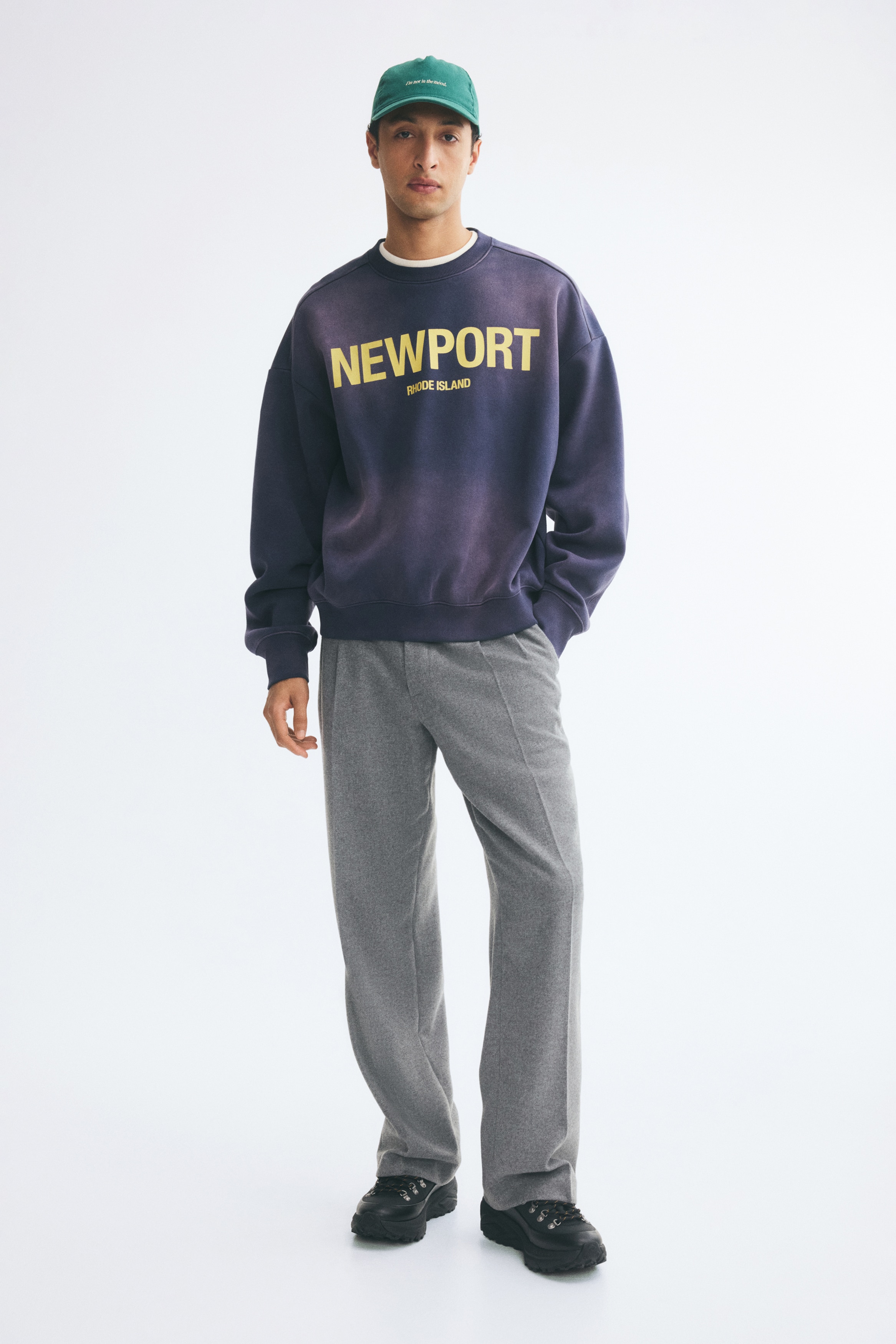 Grey trousers are shown from the front, featuring a structured waistband with belt loops and a concealed front fastening. The design displays single front pleats and sharp pressed creases running down the centre of the straight, wide legs.