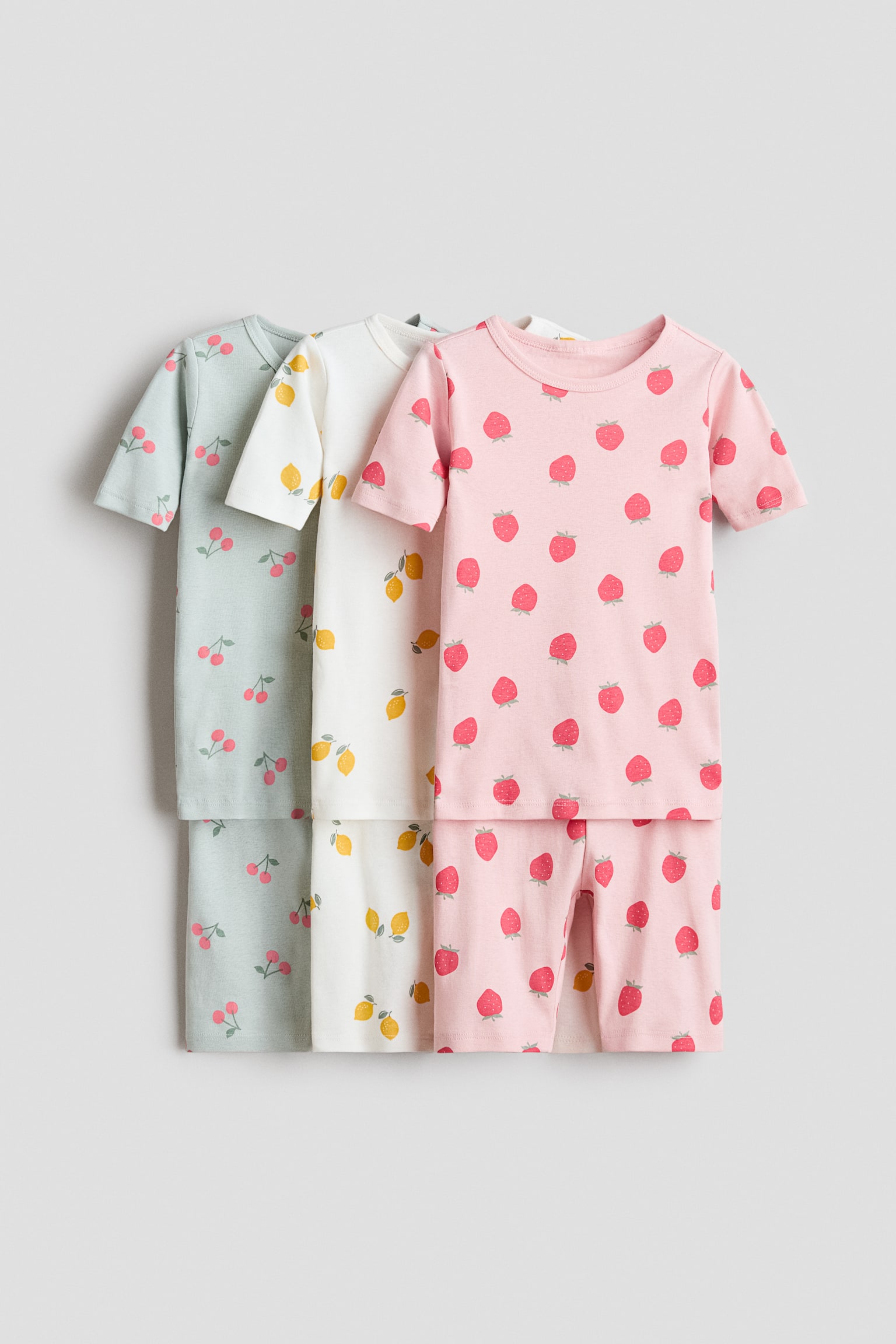 Three children's pajama sets are displayed from the front, each featuring a short-sleeved top and matching shorts. The first set is light green with red cherry prints, the second is white with yellow lemon prints, and the third is pink with red strawberry prints.