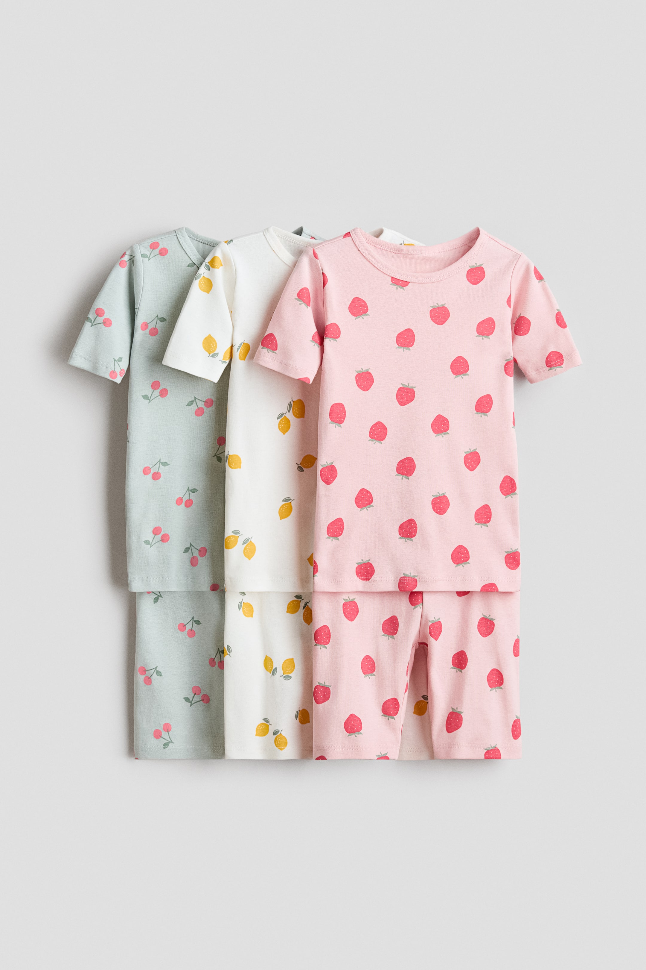 View larger image: Three children's pajama sets are displayed from the front, each featuring a short-sleeved top and matching shorts. The first set is light green with red cherry prints, the second is white with yellow lemon prints, and the third is pink with red strawberry prints.