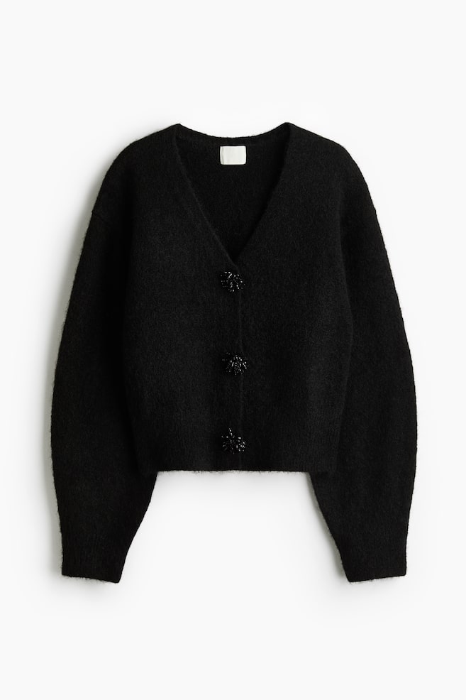 A black knit cardigan is facing front, showcasing a fuzzy texture, a V-neckline, and long voluminous sleeves with ribbed cuffs. The cropped cardigan is fastening down the center with three large, embellished black buttons. - 1