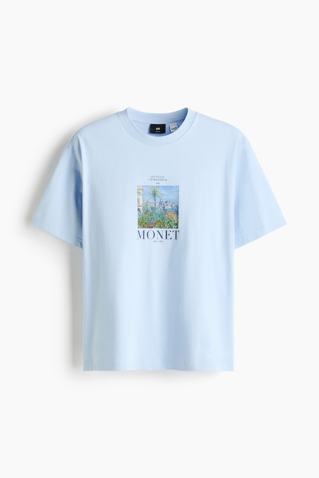 A light blue t-shirt is facing forward, featuring a ribbed crew neck and short sleeves. A rectangular graphic print is centered on the chest, depicting a colorful landscape scene with buildings, palm trees, and lush greenery, with text above and below the scene reading 'LES VILLAS A BORDIGHERA 1884' and 'MONET 1840 - 1926'. - 1