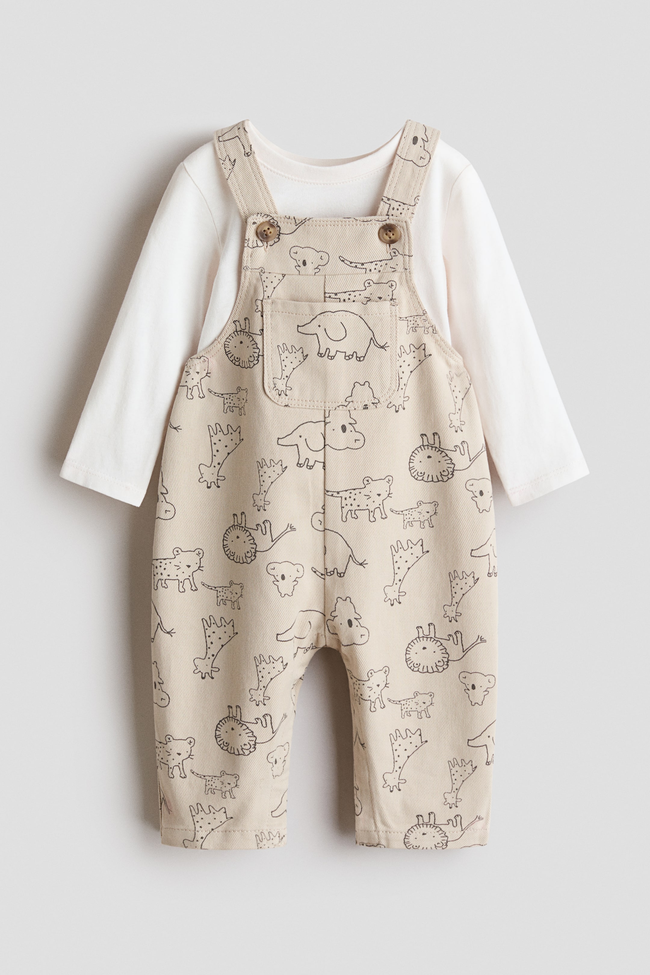 View larger image: A baby’s ensemble is showcased front-on, featuring beige overalls adorned with black animal line illustrations, layered over an off-white long-sleeved top. The overalls boast adjustable shoulder straps secured with brown buttons at the bib and a central patch pocket.