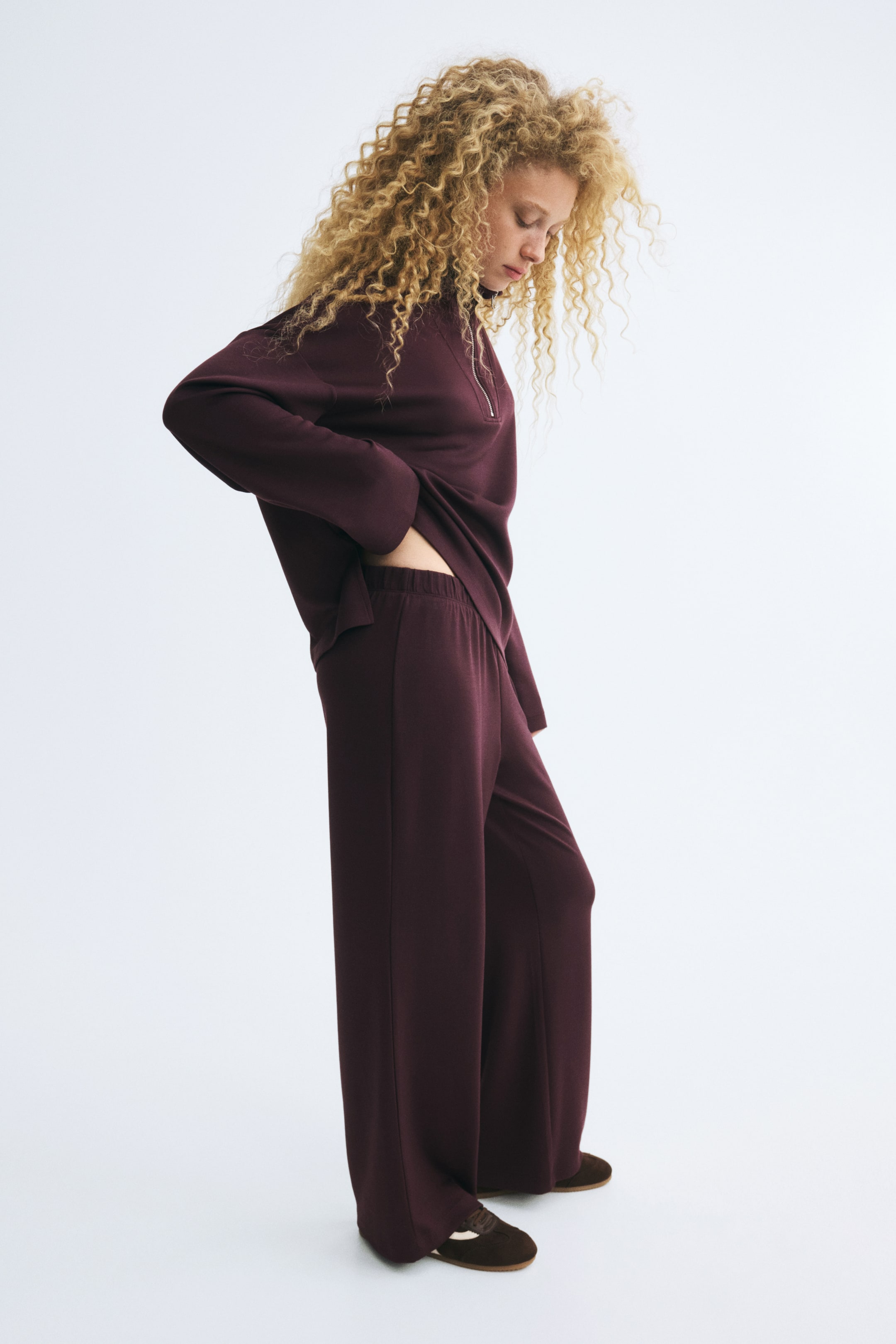 Größeres Bild ansehen: Ein bordeauxfarbenes, aufeinander abgestimmtes Set aus Half-Zip-Pullover und Hose mit weitem Bein wird von dem Model getragen. Das Model steht in einer seitlichen Profilansicht, blickt nach unten und ruht mit einer Hand nahe dem elastischen Bund der Hose, während es den Saum des locker geschnittenen Oberteils leicht anhebt.