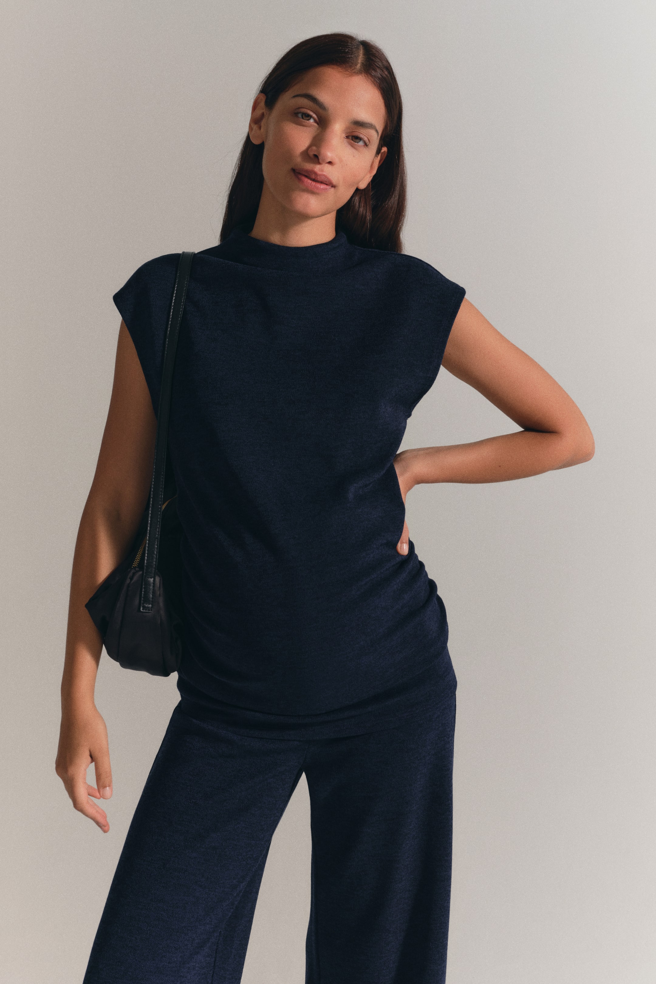 Voir une image plus grande: L’ensemble assorti en jersey bleu foncé comprend un haut à col montant et manches courtes avec coutures latérales froncées et un pantalon à jambe droite, porté par le mannequin. Le mannequin est debout, une main appuyée sur la hanche, tout en portant un sac à bandoulière noir.