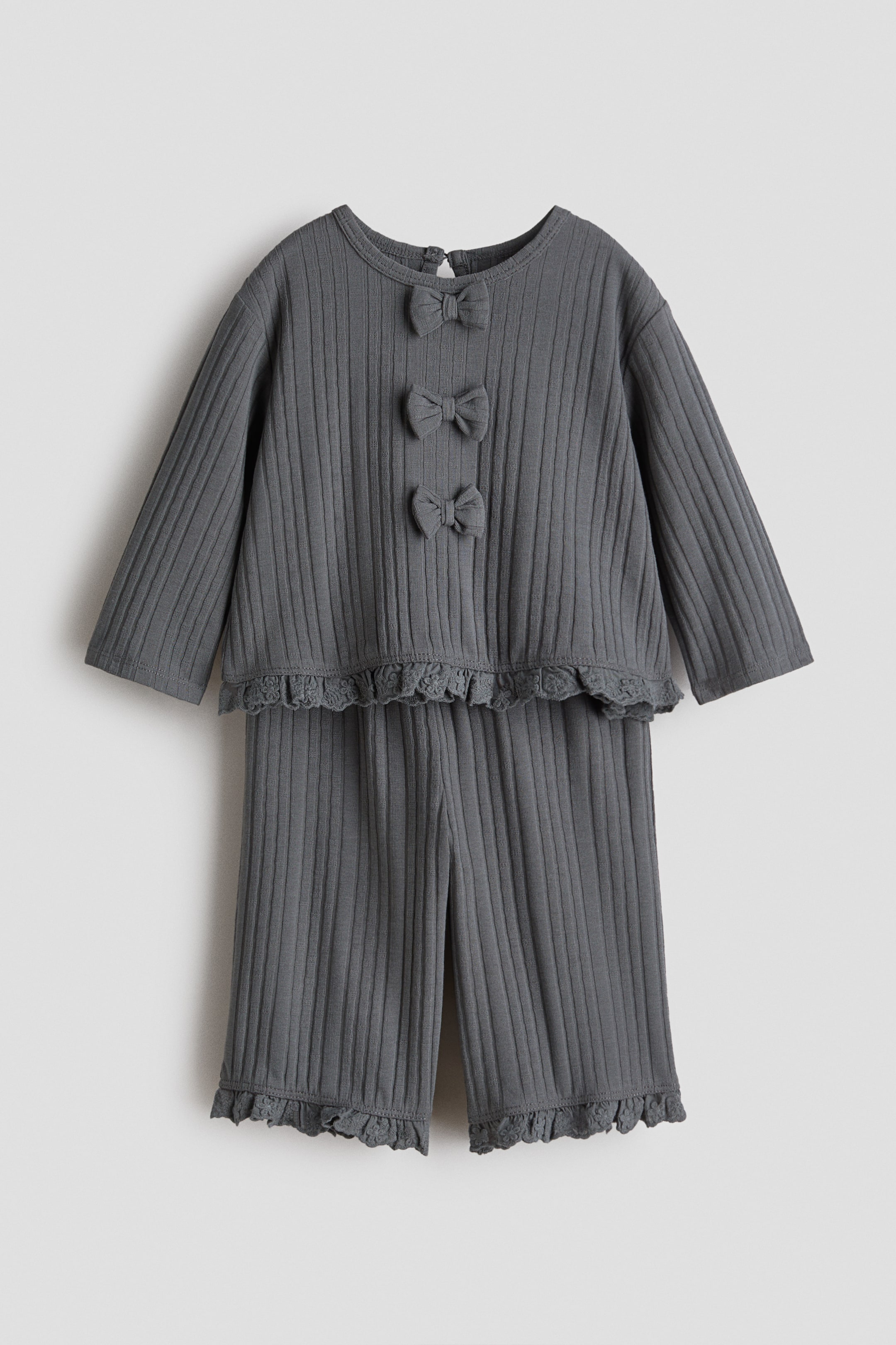 Ingrandisci l'immagine: Un set due pezzi grigio scuro a coste presenta un top a maniche lunghe con scollo rotondo, tre fiocchi decorativi sul davanti e un bordo in pizzo arricciato lungo l'orlo. I pantaloni lunghi coordinati mostrano lo stesso bordo in pizzo arricciato ai polsini.