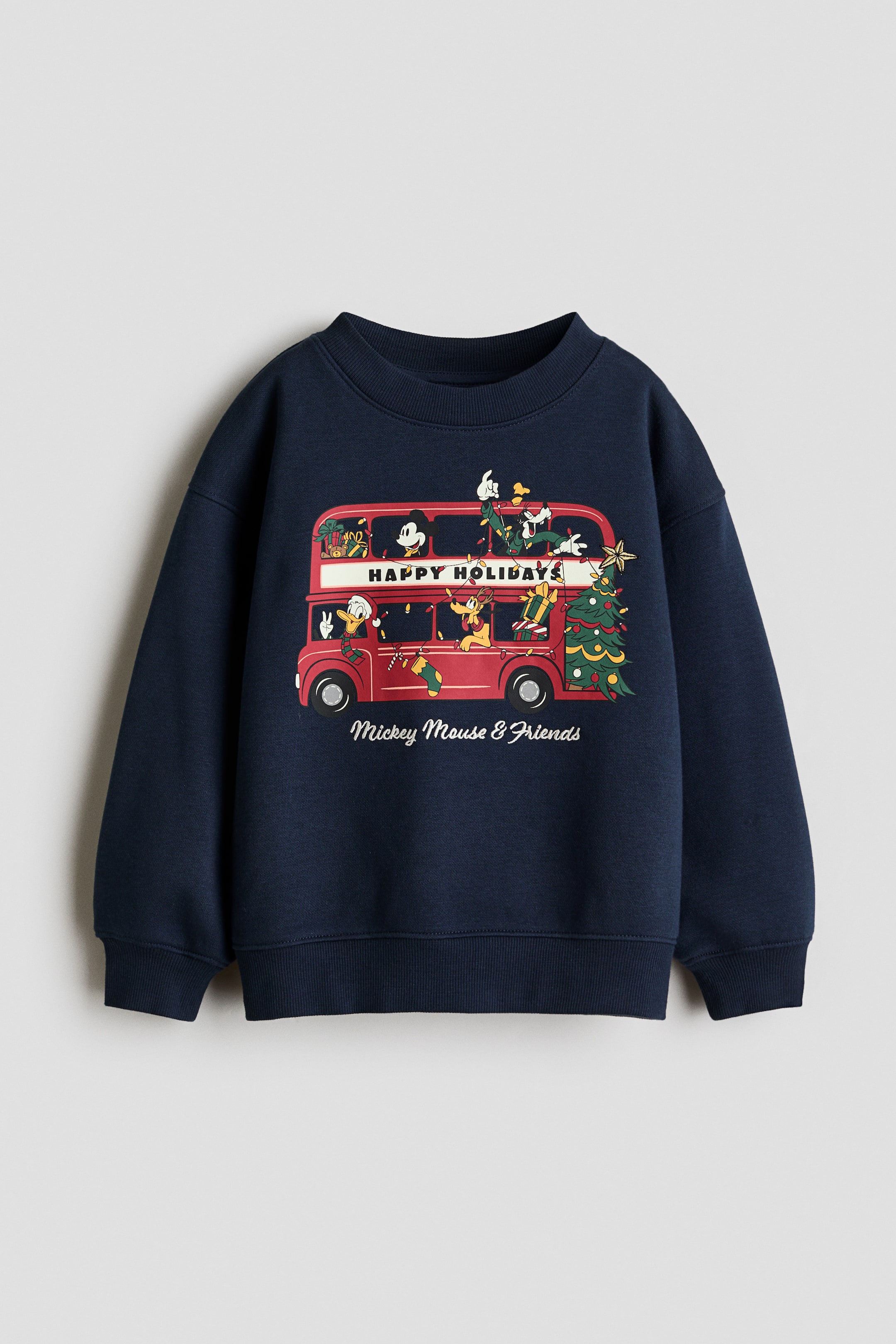 View larger image: This front-facing view shows a navy blue, loose-fit sweatshirt featuring a round, rib-trimmed neckline, dropped shoulders, and long sleeves with ribbed cuffs and a ribbed hem. The front displays a colorful printed graphic of Disney characters on a red double-decker bus decorated for the holidays, with the text "HAPPY HOLIDAYS" and "Mickey Mouse & Friends" printed below.