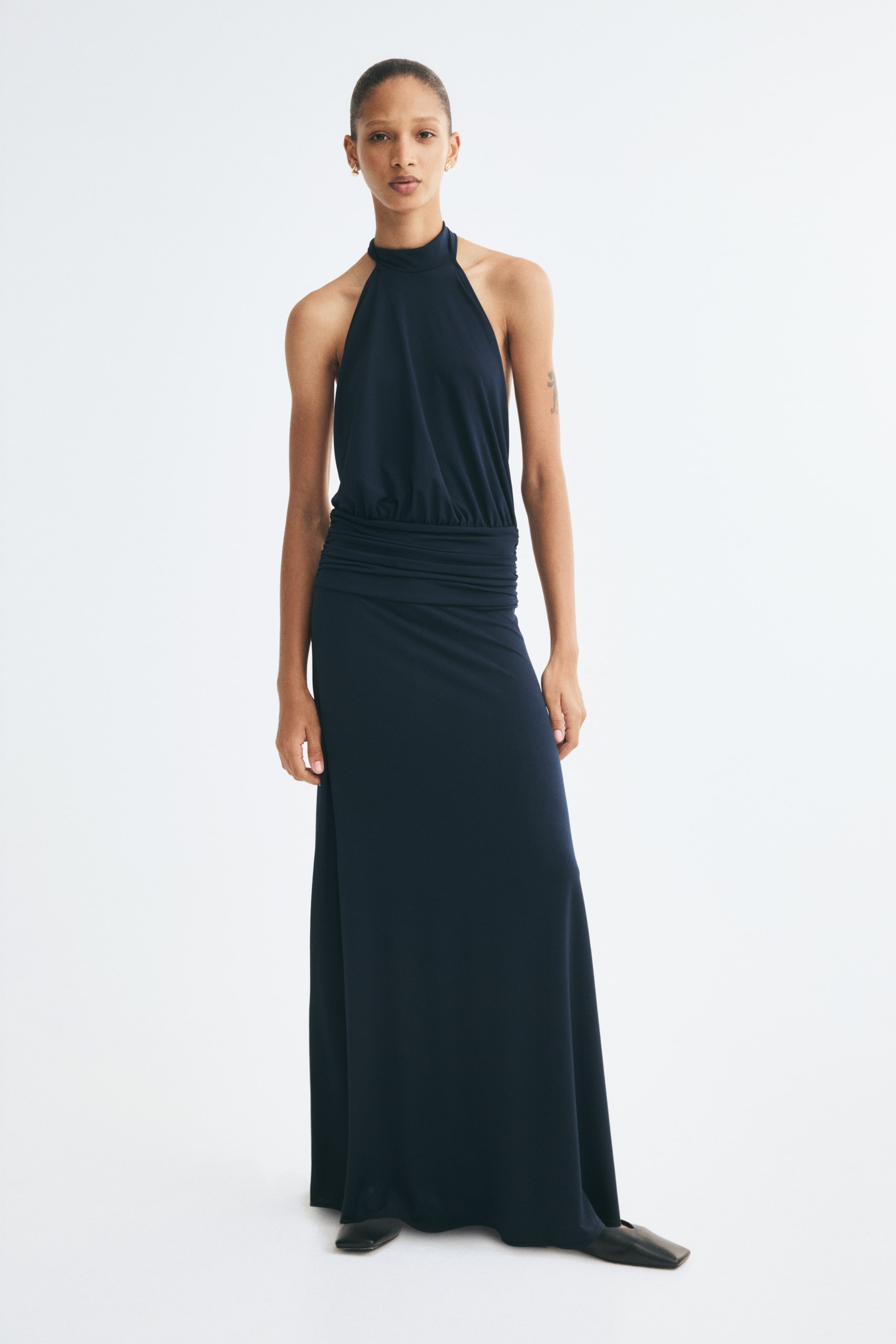 Se større billede: En mørk marineblå stretch-jersey maxi-kjole ses forfra med halterneck-inspireret top, høj opretstående krave og et smalt ærmeløst snit. Kjolen er designet med rynkede, draperede detaljer hen over taljen og falder ned i en lang, lige maxinederdel.
