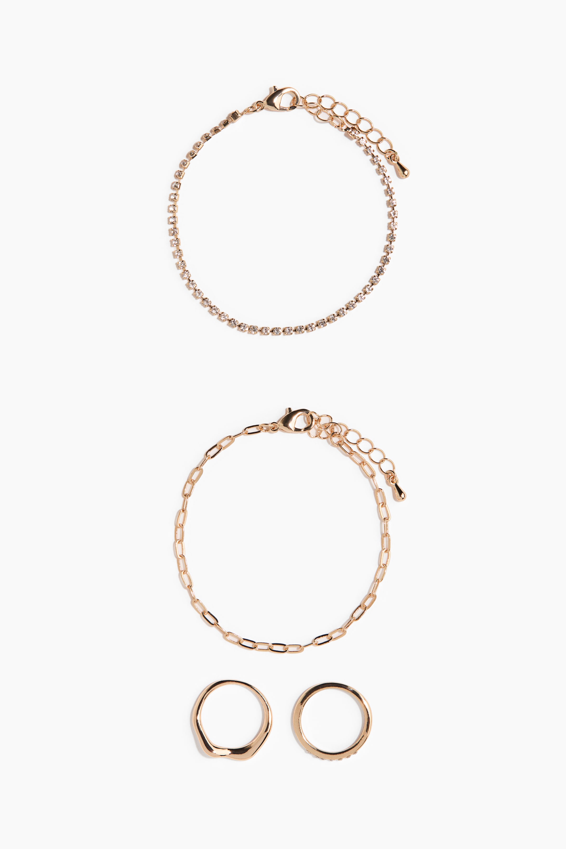 Visualizza immagine più grande: Questo set di gioielli color oro presenta due bracciali e due anelli disposti in una flat lay frontale. Il bracciale superiore mostra una delicata fila di piccoli strass trasparenti, il bracciale centrale mette in evidenza una catena a maglie paperclip, mentre quello inferiore presenta due fasce color oro, tra cui una con una silhouette ondulata e organica.