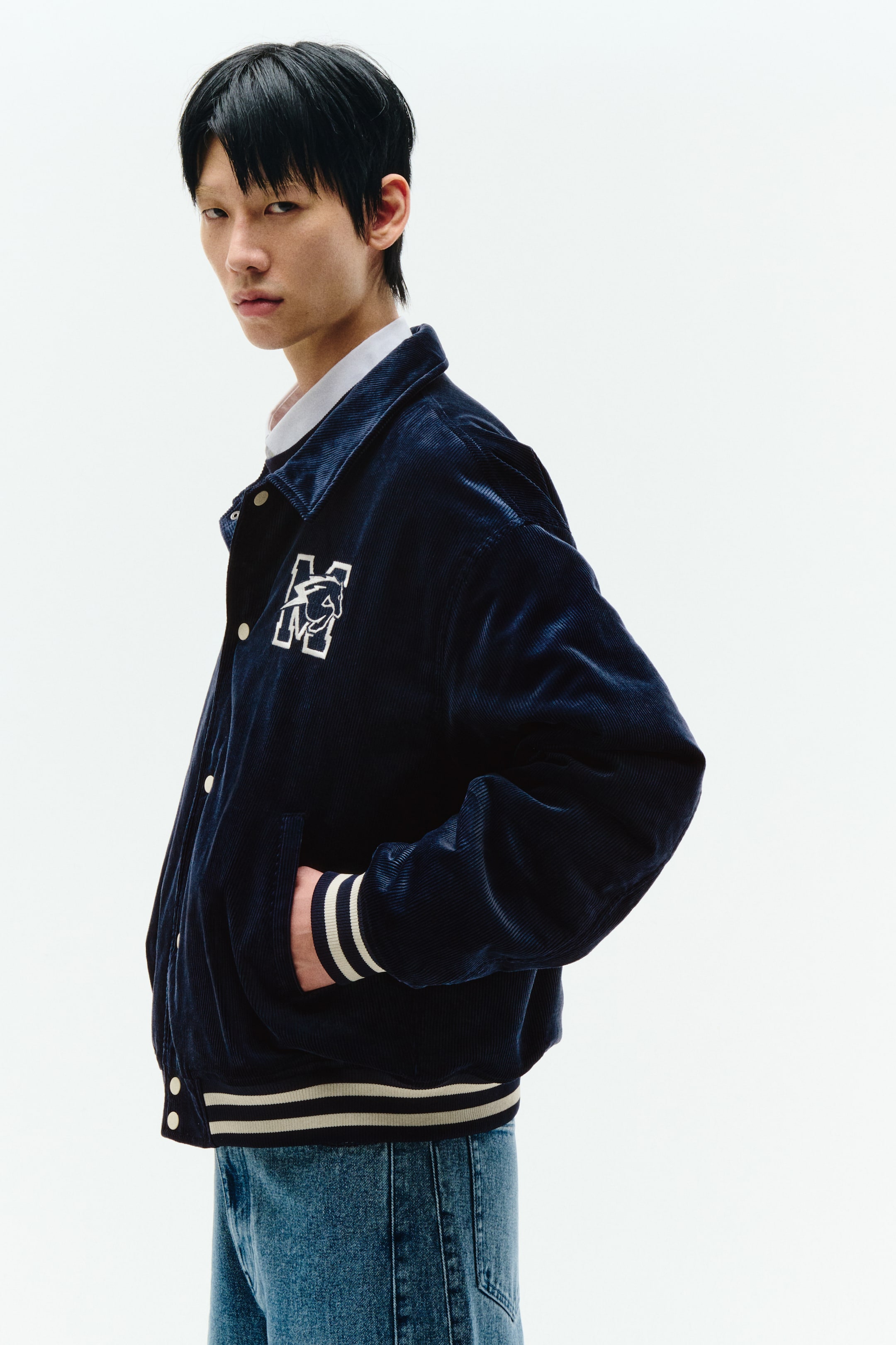 拡大画像を表示: ネイビーのコーデュロイのバーシティジャケットが、左胸に動物のデザインが施された白い刺繍の「M」を特徴とし、三分の四の角度から見せられています。尖った襟は白いシャツの上に重ねられ、前面にはスナップボタンが閉じ、ストライプのリブ付き袖口と裾が付いています。ポケットに手を入れています。