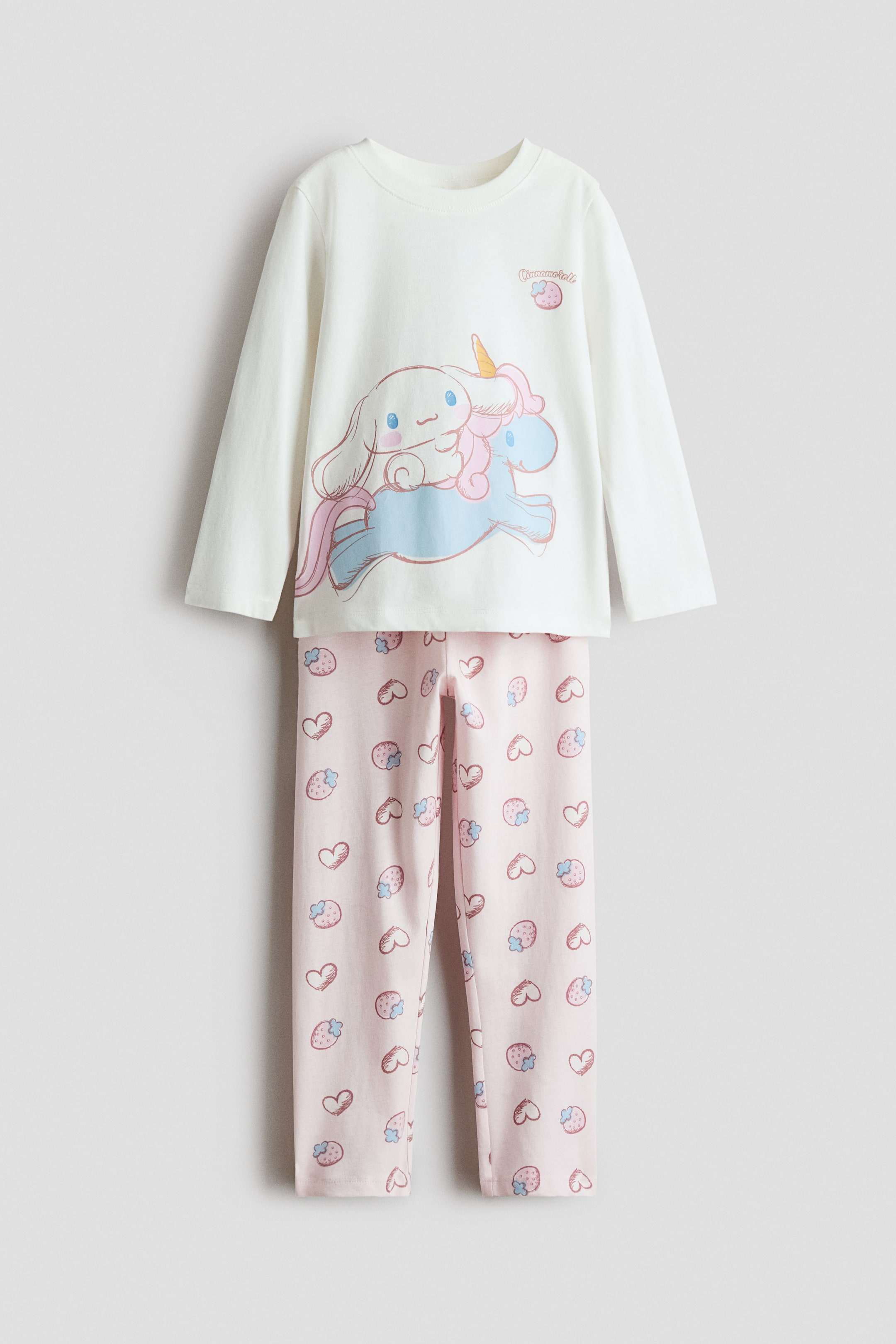 View larger image: A front-facing pyjama set displays a white, long-sleeved top with a rib-trimmed round neckline and a large cartoon graphic at the front. The matching light pink pants feature an elasticated waist and an all-over print of hearts and strawberries.