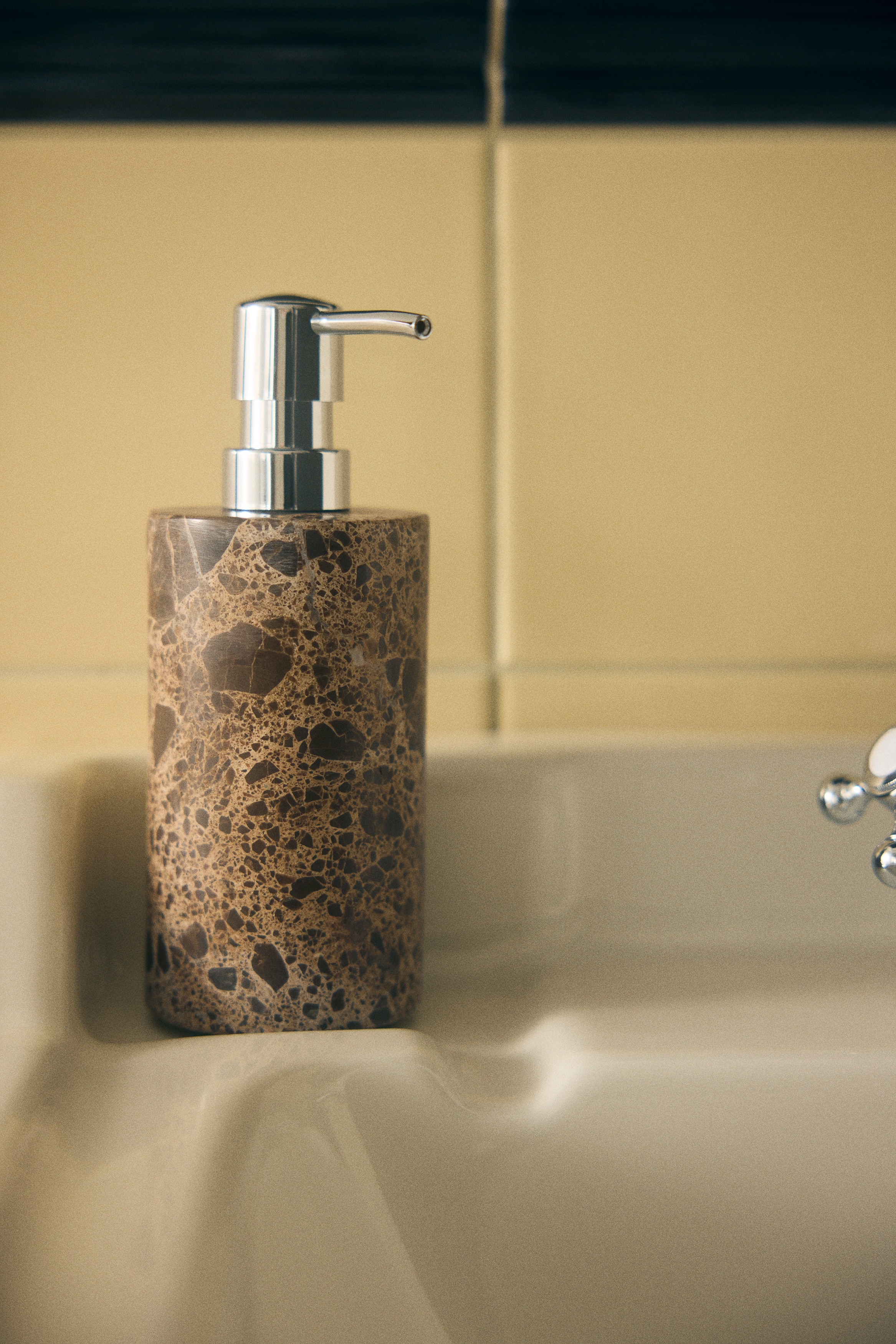 Un distributore di sapone cilindrico presenta un corpo in marmo marrone con tonalità terrose variegate e motivi screziati più chiari. Un meccanismo a pompa in cromo lucido copre la parte superiore, con il suo beccuccio che si estende orizzontalmente in avanti.