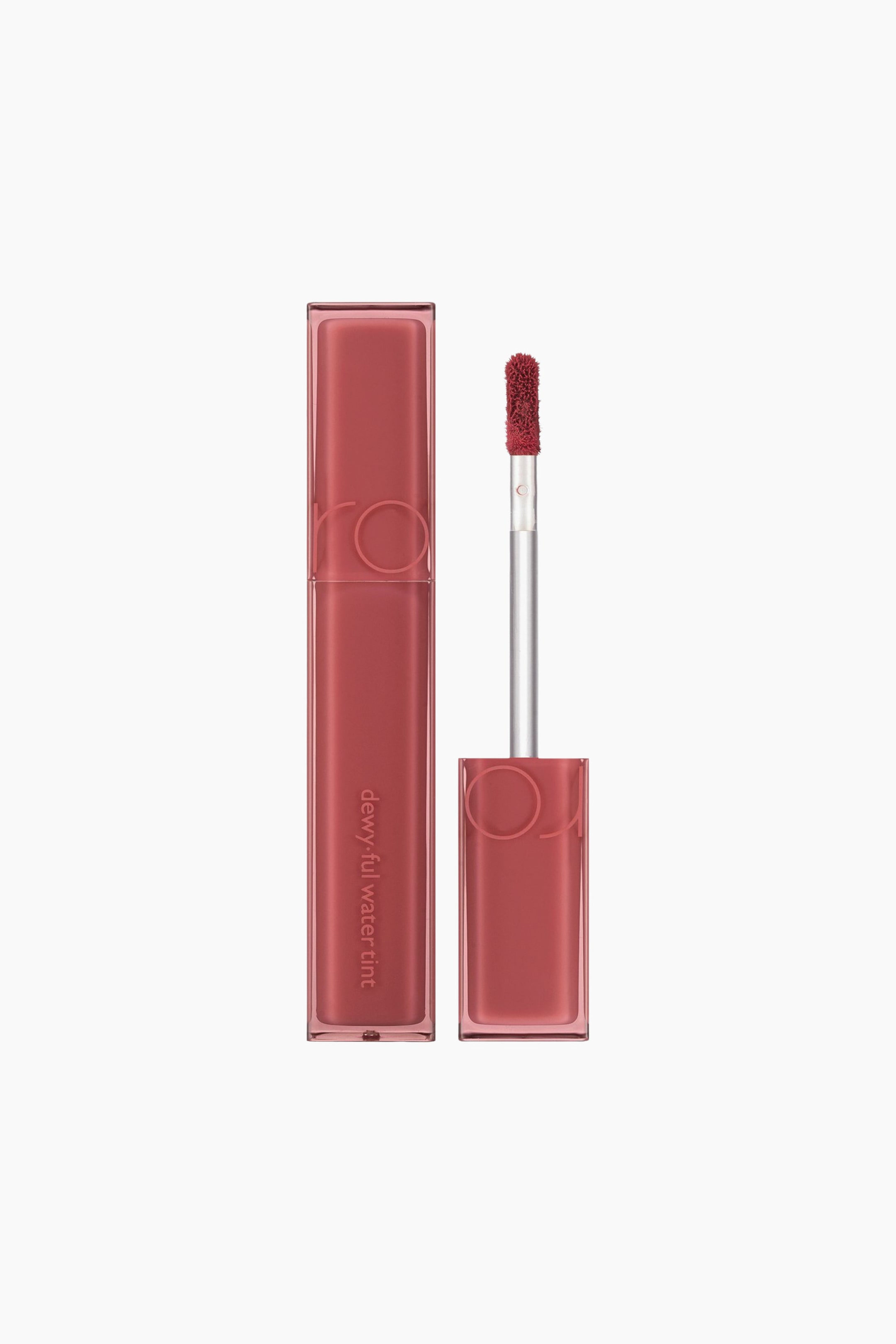 Vis større bilde: Et rektangulært lip tint-rør, merket «rom&nd dewy-ful water tint», står oppreist og er fylt med en rødbrun væske. Ved siden av vises en applikator med en flocket doe-foot-tupp, dekket av samme tint, mens den matchende rødbrune hetten ligger under tuppen.