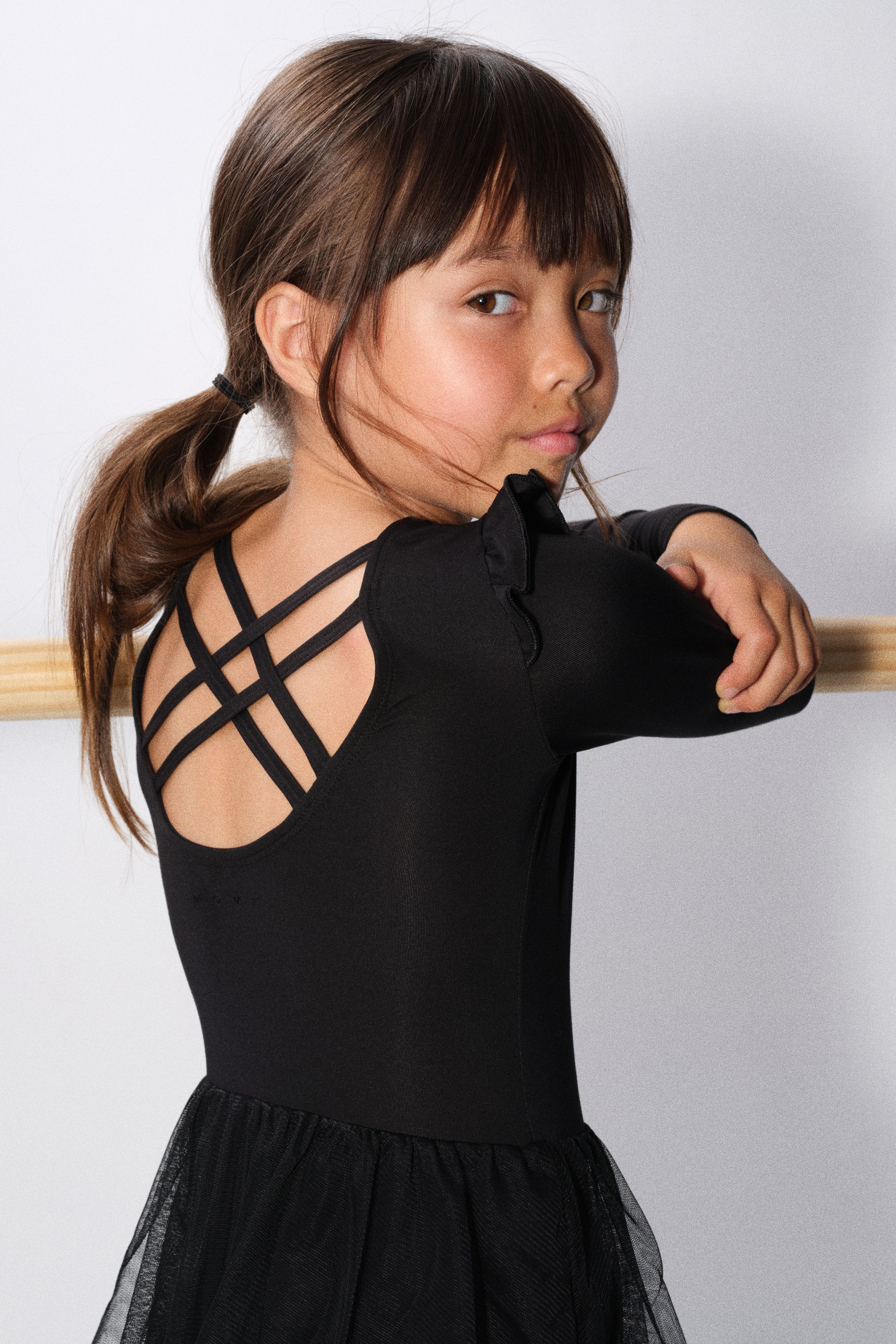 A black long-sleeved dance leotard is shown from the front, featuring ruffle details at the shoulders and a gathered sweetheart neckline. It has visible crisscross back straps and an attached sheer black tulle skirt layered over the leotard bottom.
