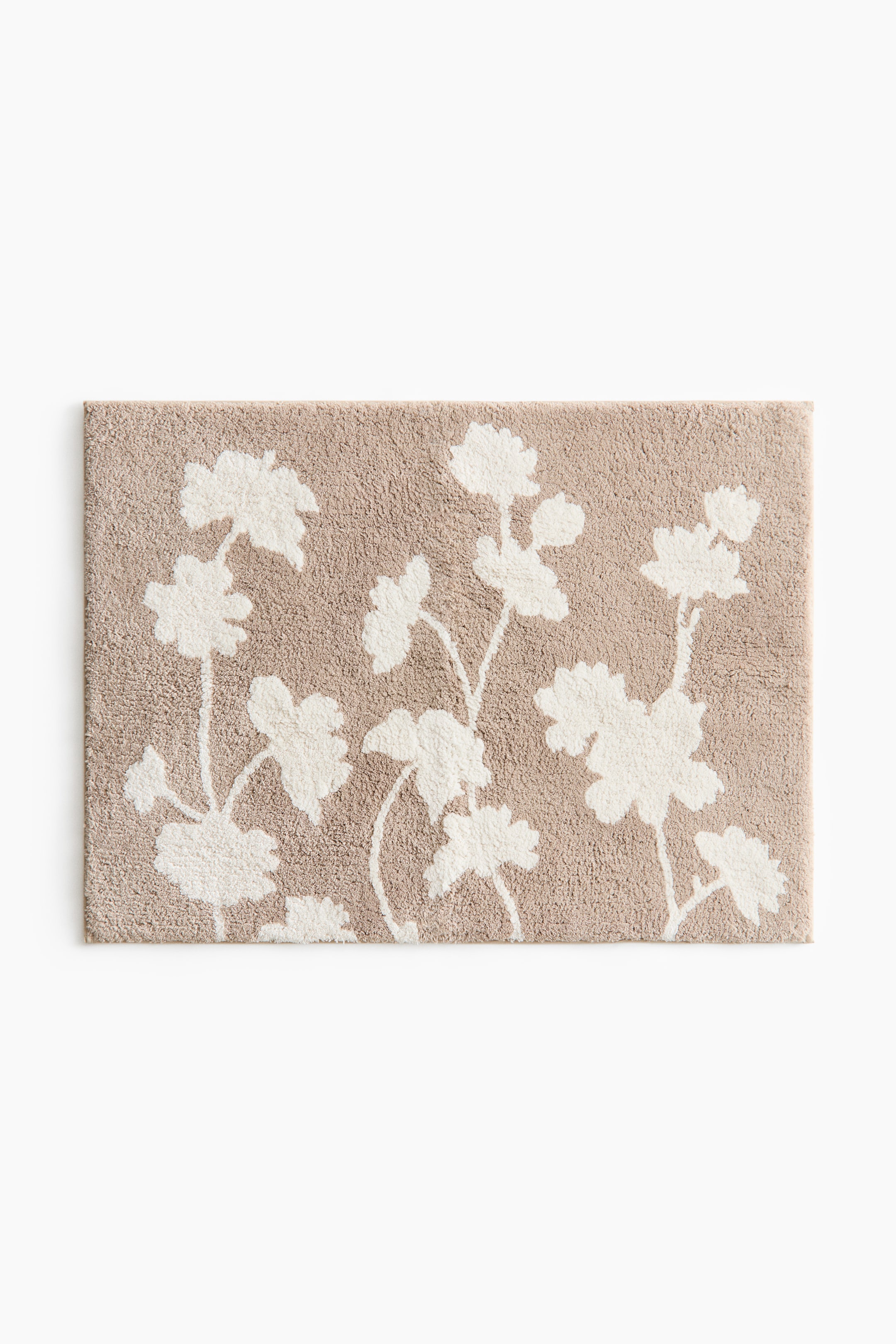 View larger image: The bath mat is rectangular and showcases a tufted light brown surface adorned with a white floral and leaf pattern throughout. It features stylized blossoms and foliage intricately woven across the front.