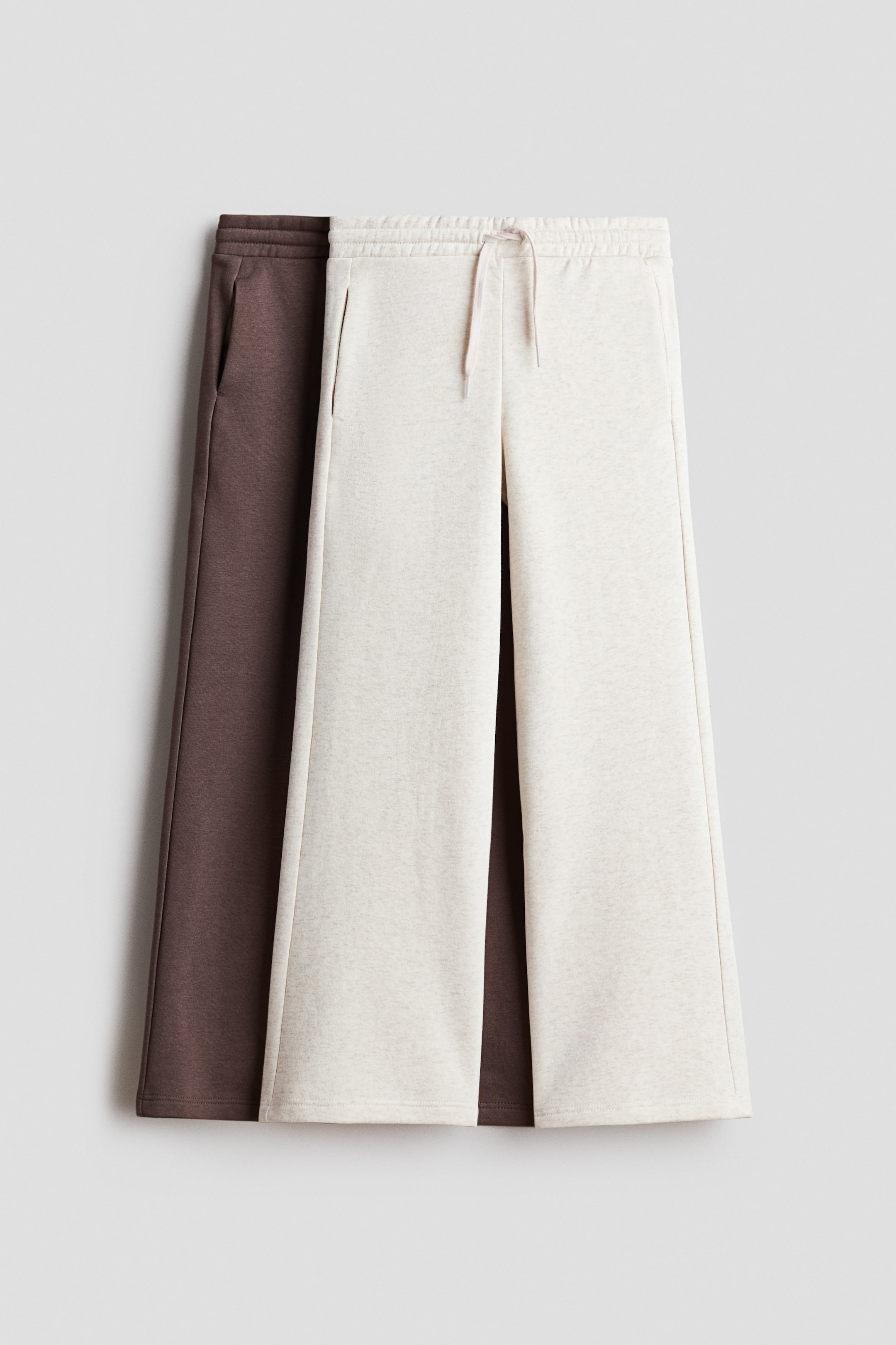 View larger image: Two pairs of wide-leg sweatpants are showcased, with the front light beige pair exhibiting an elasticated drawstring waist and side pockets. This pair is partially overlapping a brown pair, which features an elasticated waistband and side pockets.