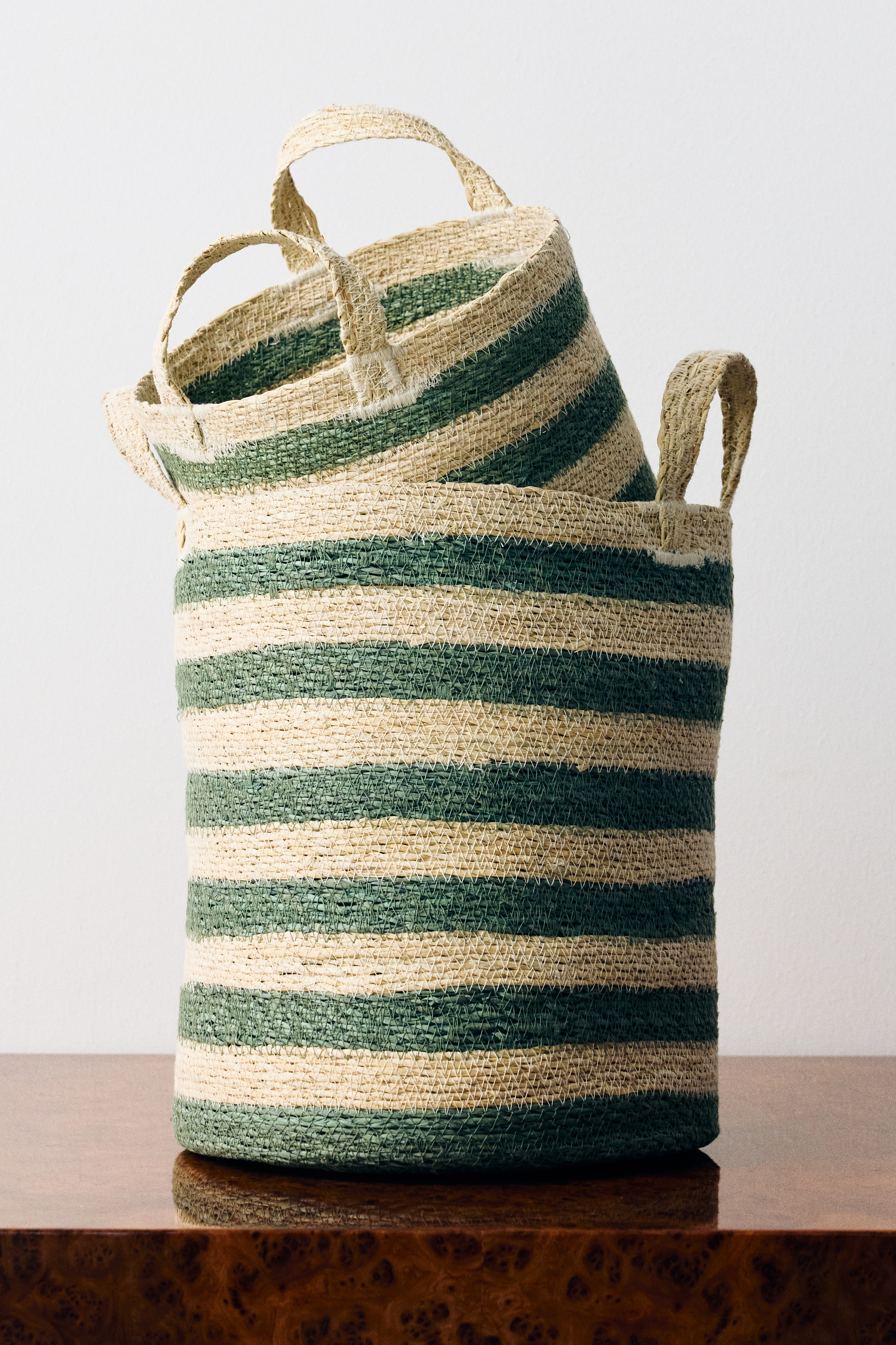 A woven straw basket is shown in a close-up view, featuring alternating horizontal stripes of natural beige and sage green. The basket is displaying a thick, arched woven handle attached to the rim with visible white stitching that reinforces the textured weave.