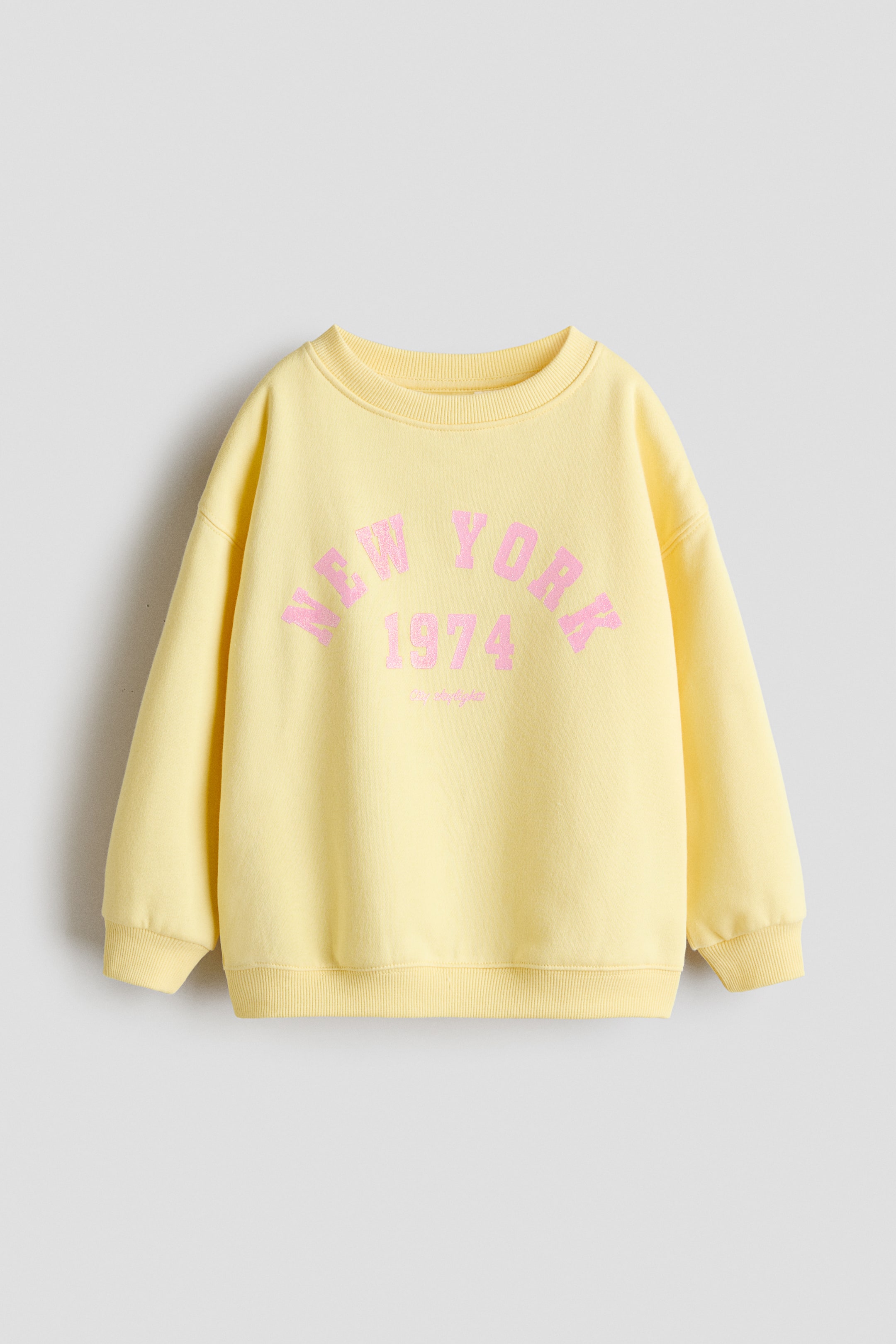 View larger image: A yellow sweatshirt, made from printed sweatshirt fabric, is facing FRONT. It is featuring 'NEW YORK 1974' in pink arched text on the chest, a round rib-trimmed neckline, gently dropped shoulders, and ribbing at the cuffs and hem.