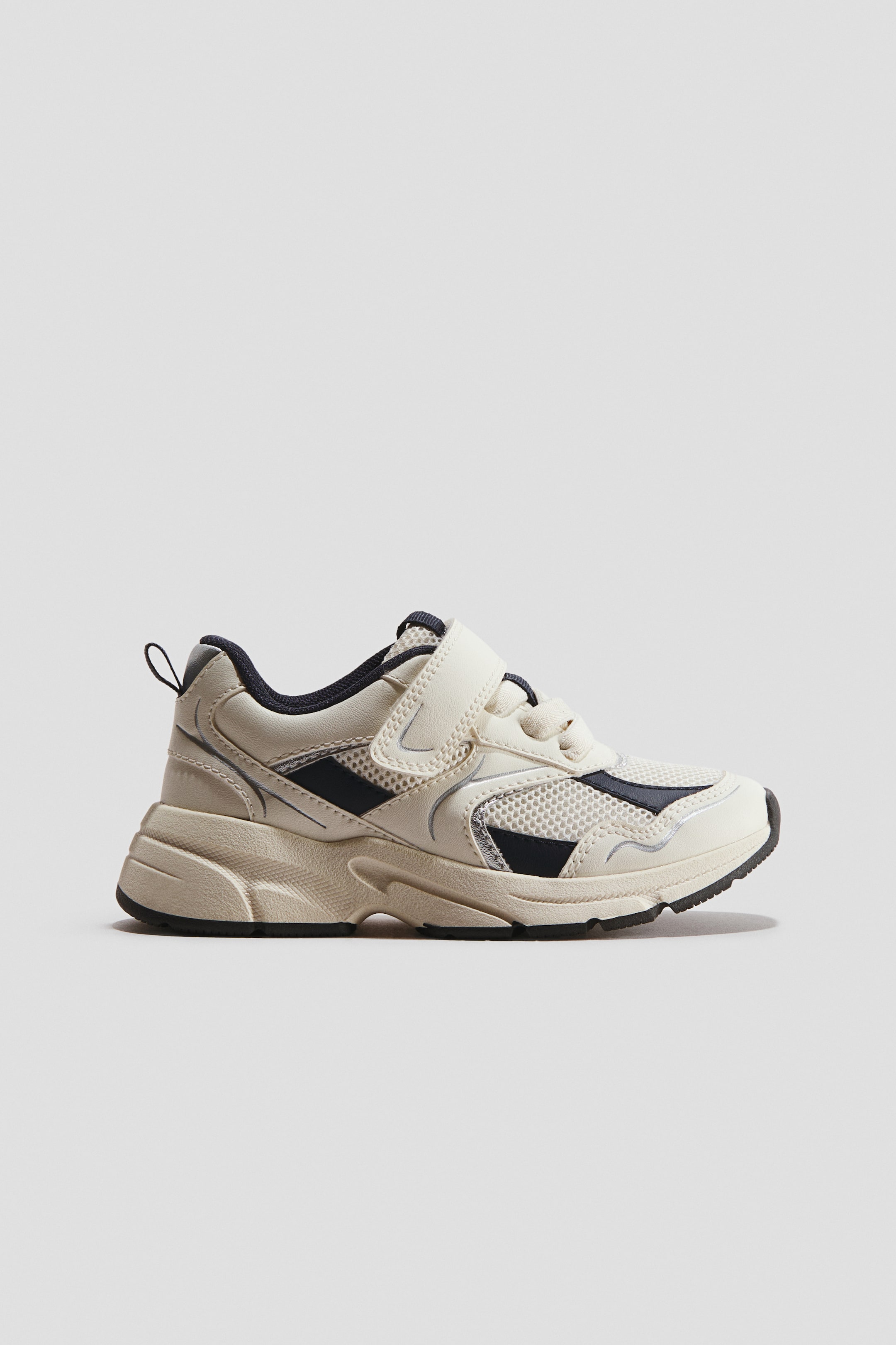 View larger image: An off-white trainer is facing left, showcasing mesh panels, navy and silver-toned synthetic overlays, a chunky textured sole, and a Velcro strap securing elastic laces.