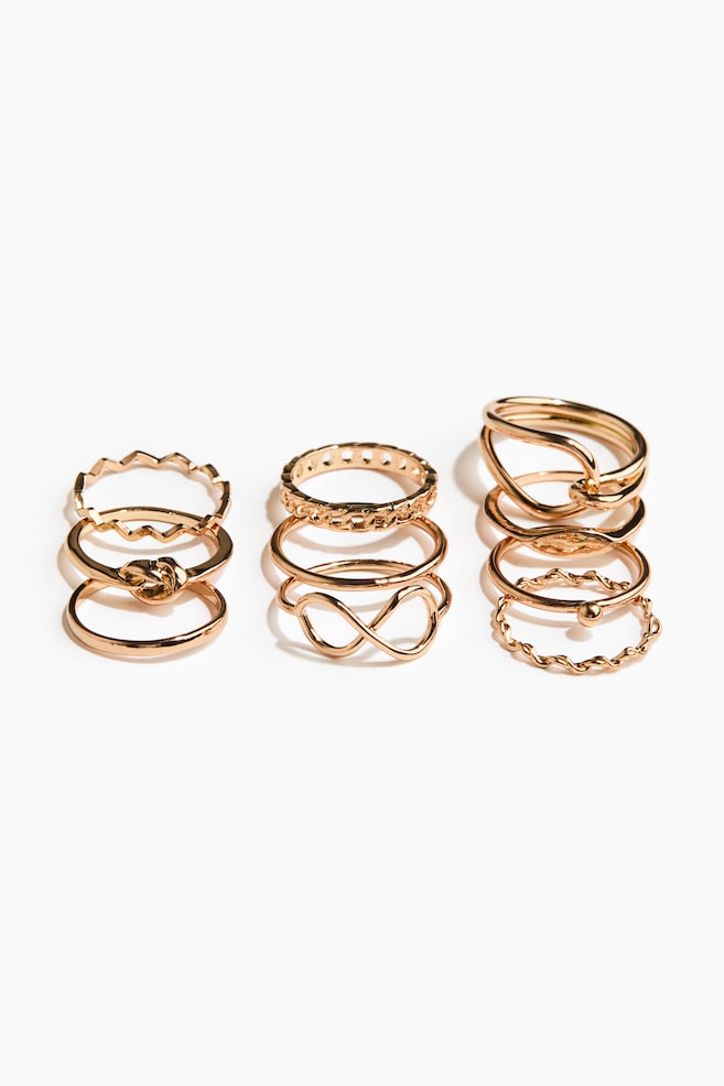 A set of ten gold-toned rings is being displayed, showcasing various designs. The rings are featuring a zig-zag patterned band, a knot detail, a chain-link pattern, an infinity symbol, two intertwined bands, a small spherical bead, and several plain, wavy, and twisted band textures. - 1