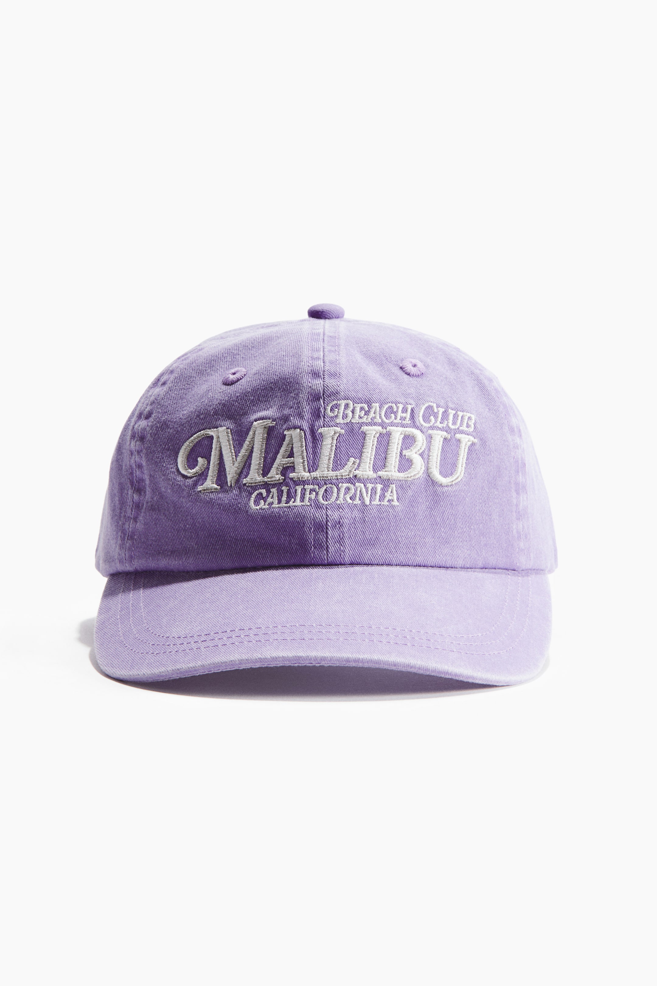 View larger image: A lavender cotton twill cap is facing forward, showcasing white embroidered text that reads "BEACH CLUB MALIBU CALIFORNIA" on its crown. The cap features a washed look and a curved brim with visible stitching.