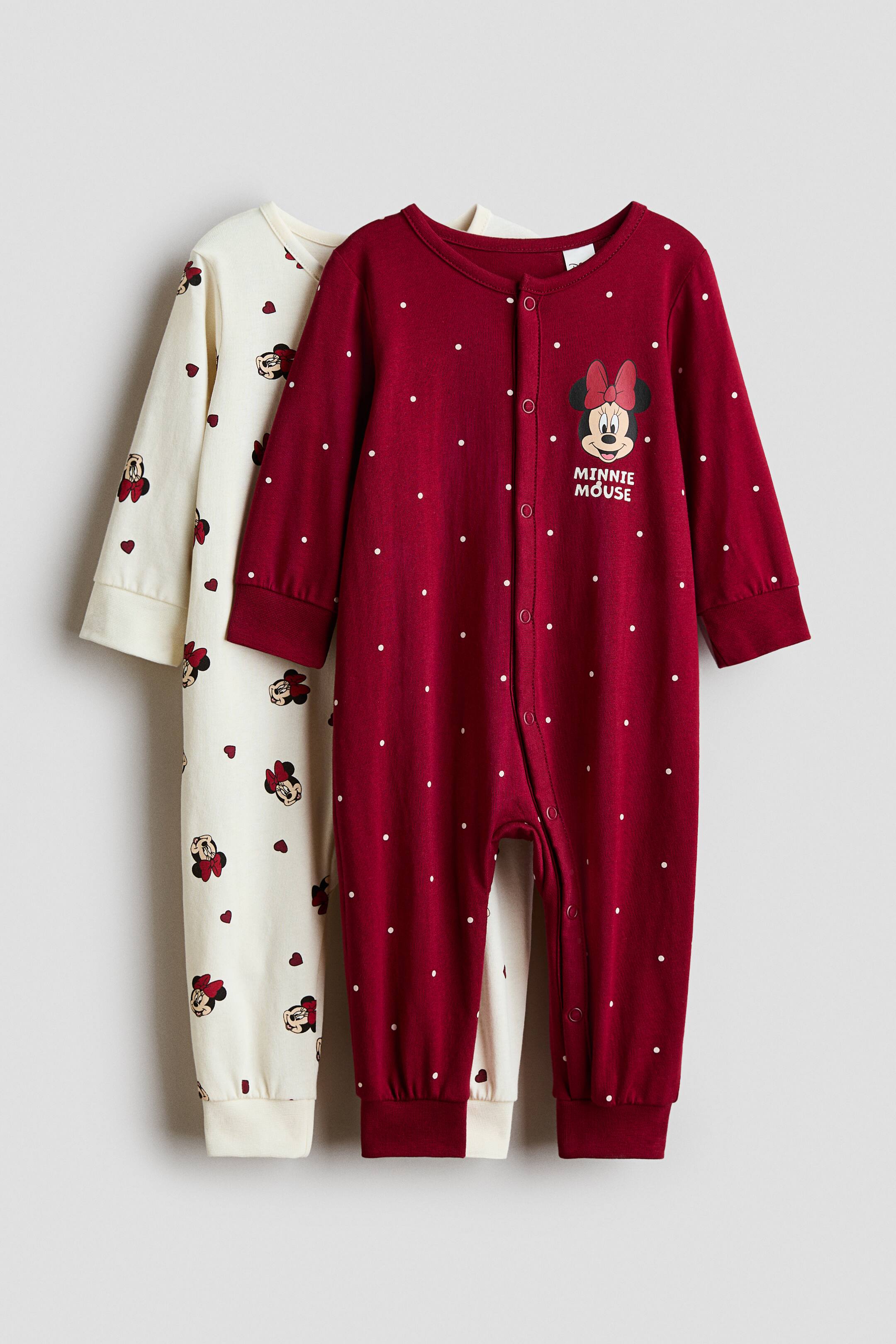 View larger image: Two cotton jersey sleepsuits are displayed from the front, with one layered slightly behind the other. The foreground sleepsuit is dark red with white polka dots and a Minnie Mouse graphic on the chest, featuring a round neckline, press studs down the front and along one leg, and wide ribbed cuffs and hems. The background sleepsuit is off-white, featuring an all-over print of Minnie Mouse heads and tiny red hearts.