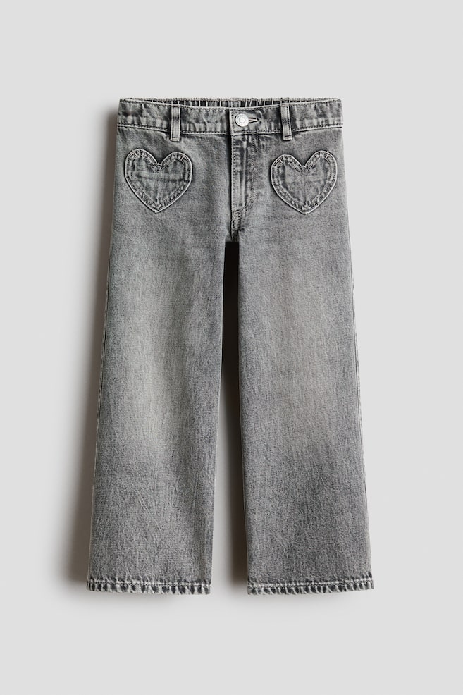 Jeans in denim di cotone grigio mostrati frontalmente, con una silhouette a gamba larga e una vestibilità morbida. I jeans presentano tasche applicate a forma di cuore sui fianchi anteriori, una cintura elastica arricciata, passanti per cintura e chiusura con bottone. - 1