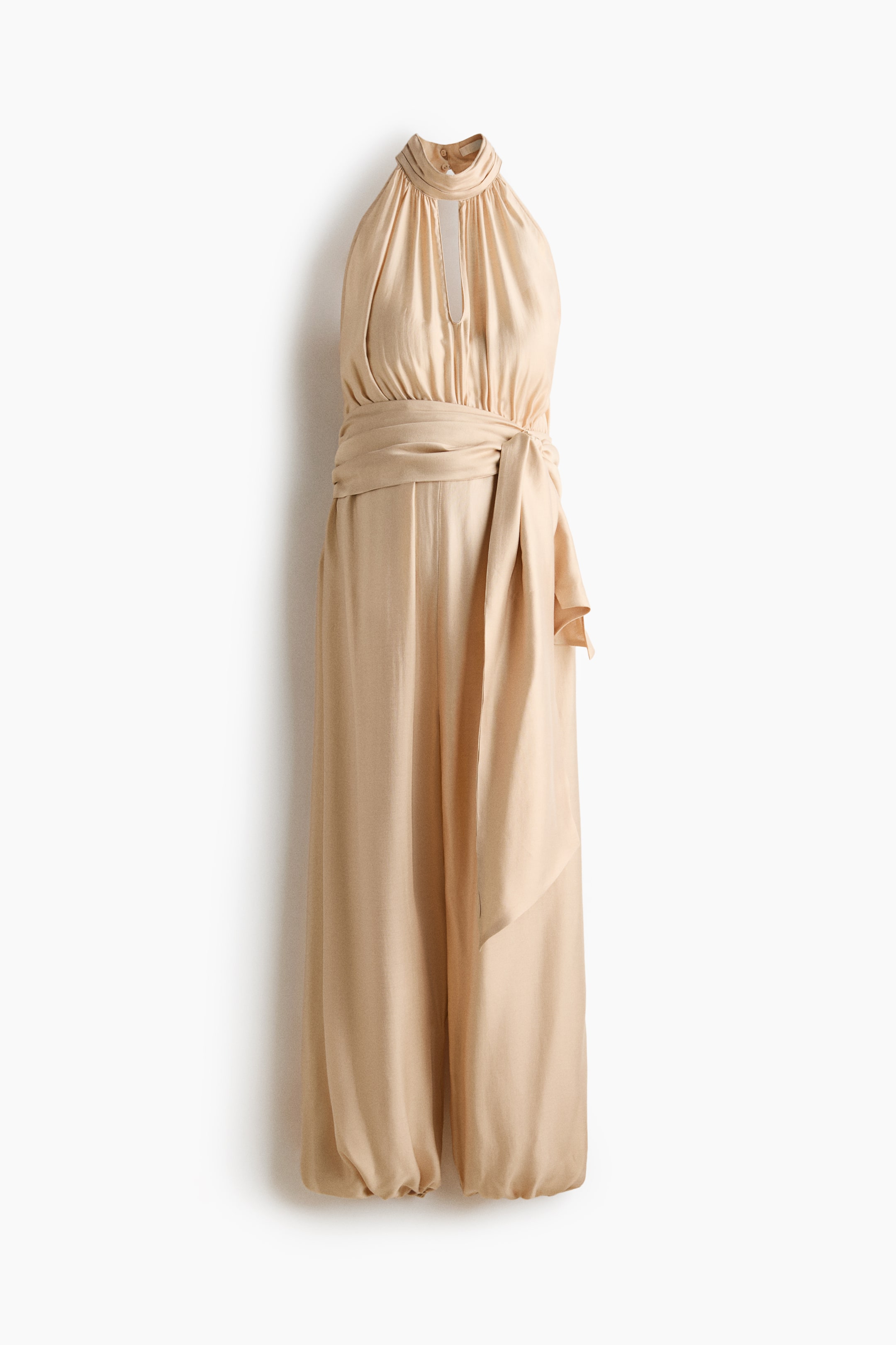 Grotere afbeelding bekijken: Een licht beige jumpsuit met een halterhalslijn en een sleutelgatuitsnijding bij de borst, en een hoge kraag die met knopen aan de achterkant van de nek wordt vastgemaakt. Een brede stoffen ceintuur accentueert de taille, en de wijde pijpen van de broek verzamelen zich bij de enkels.