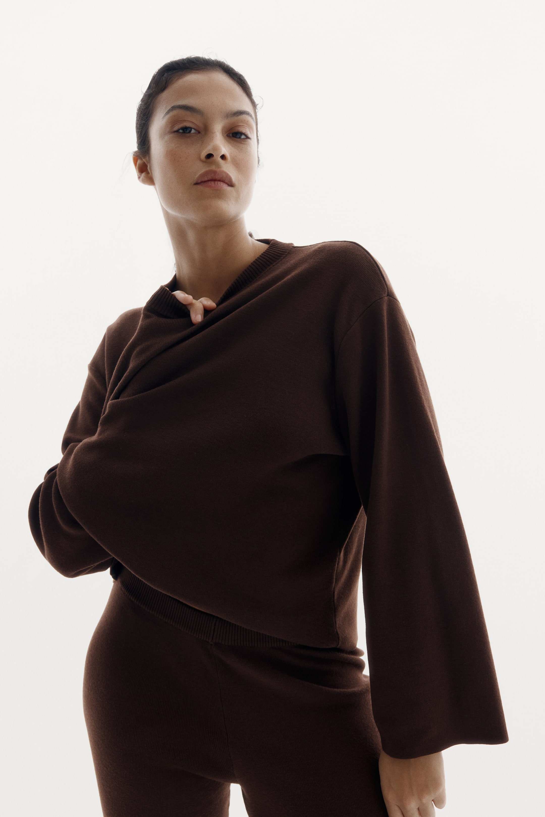 Agrandir l'image: Un haut en maille marron foncé et un pantalon assorti sont portés. Le haut présente un col rond côtelé, des manches évasées larges et un ourlet côtelé, et est conçu pour mettre en valeur son drapé ample sur un pantalon taille haute assorti qui présente une ceinture côtelée.