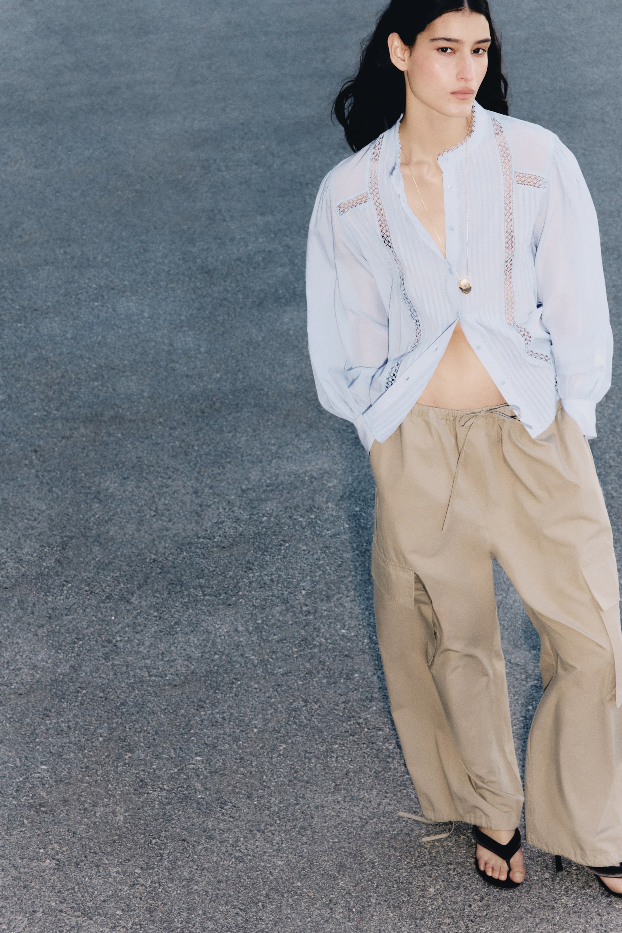 View larger image: A light blue loose-fit blouse, featuring front pintucks, lace insets, a lace-trimmed band collar, buttons down the front, and long sleeves with buttoned cuffs, is being worn by the model. The blouse is partially unbuttoned at the front, and a gold pendant necklace is resting on it.