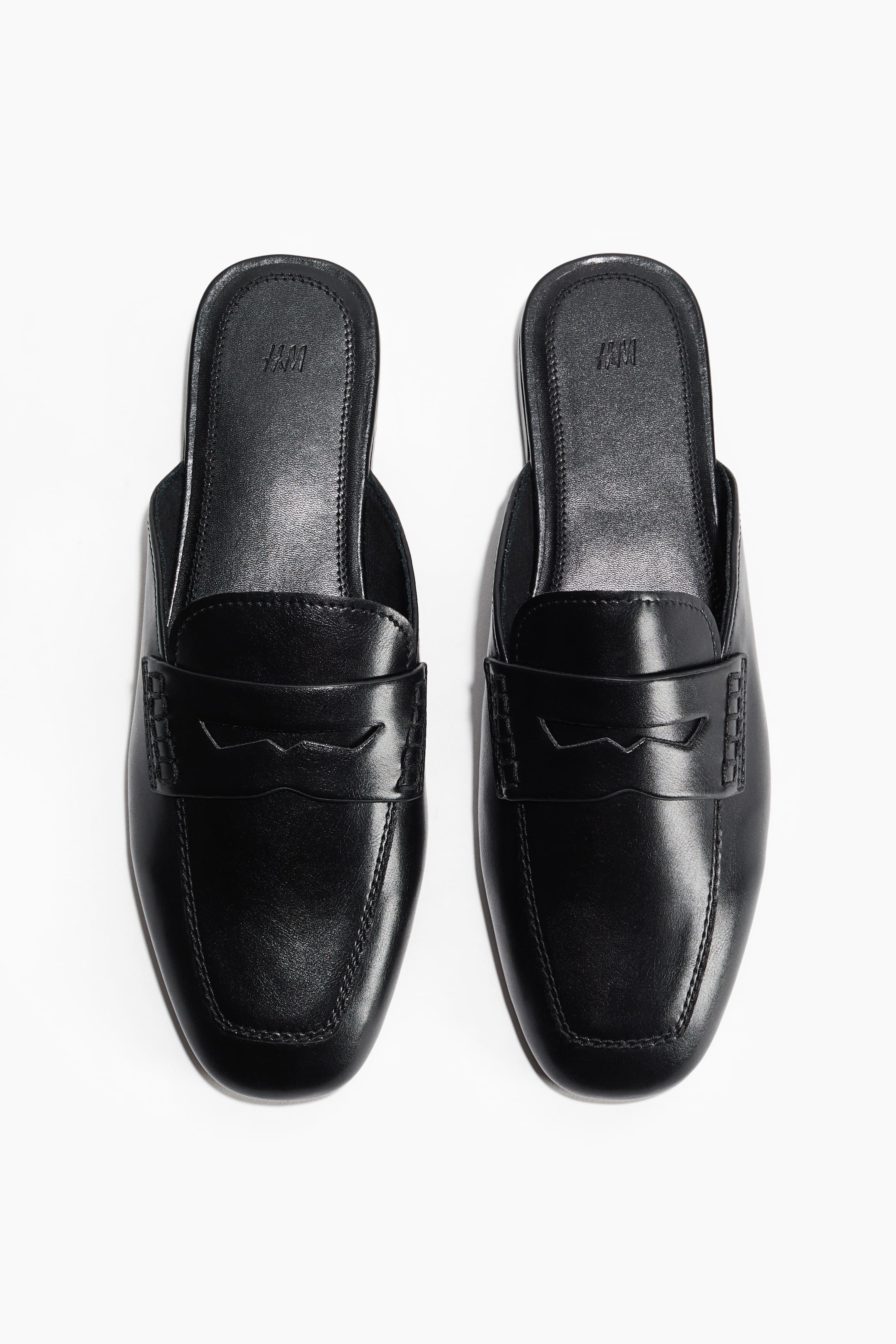 Ver imagen más grande: Dos mocasines tipo mule negros están orientados hacia adelante, cada uno con un acabado liso, una puntera cuadrada y un diseño clásico de mocasín. Una correa de montura con un recorte decorativo cruza el empeine, y las costuras visibles detallan la puntera. El logo de H&M está grabado en la plantilla negra de cada zapato.