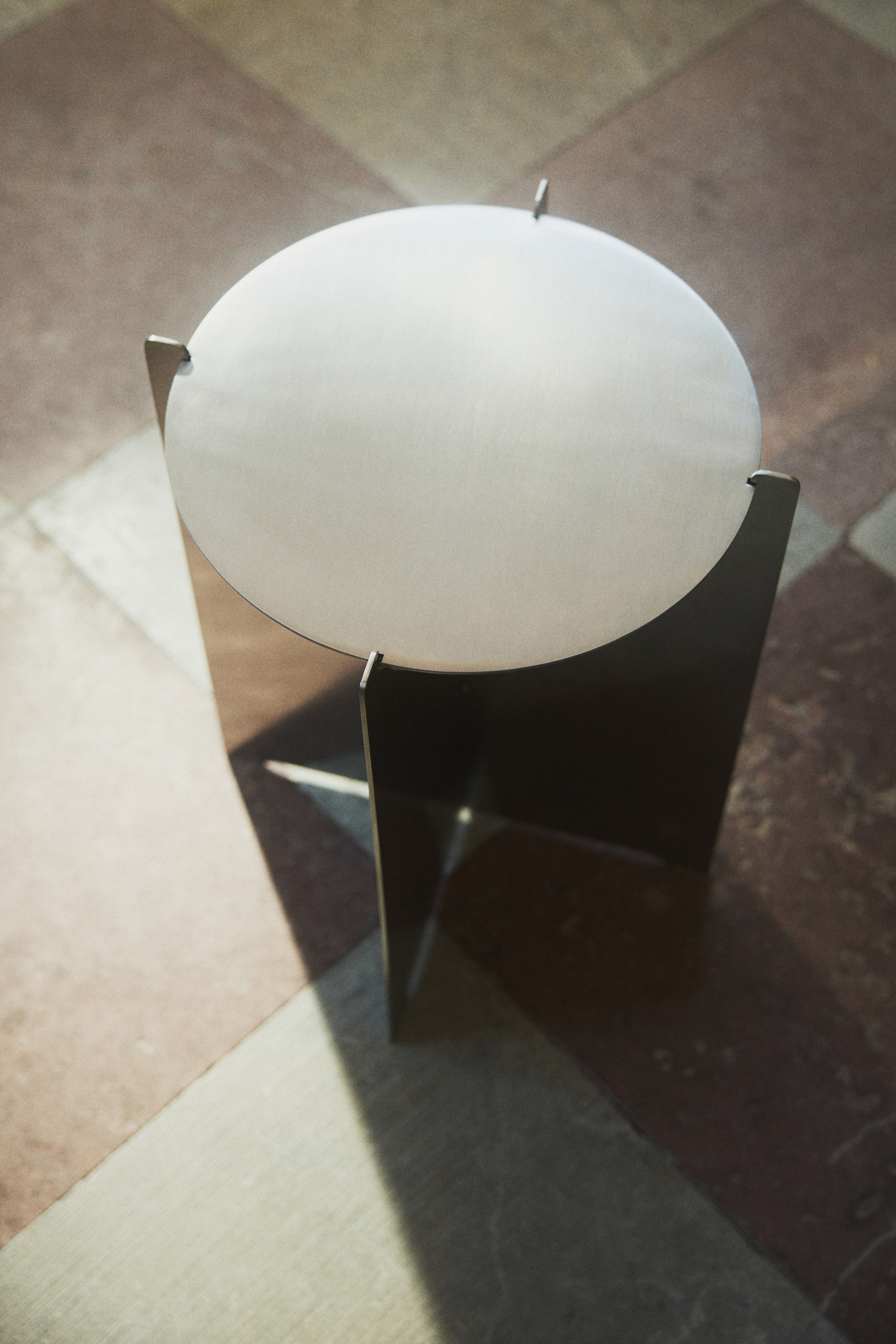 Größeres Bild ansehen: Ein hellfarbiger, runder Tisch wird aus einer hohen Perspektive betrachtet. Er verfügt über eine Tischplatte, die mit drei dunklen, flachen Paneelbeinen integriert ist. Kleine Verlängerungen der Beine ragen leicht über die Oberfläche der runden Tischplatte hinaus.