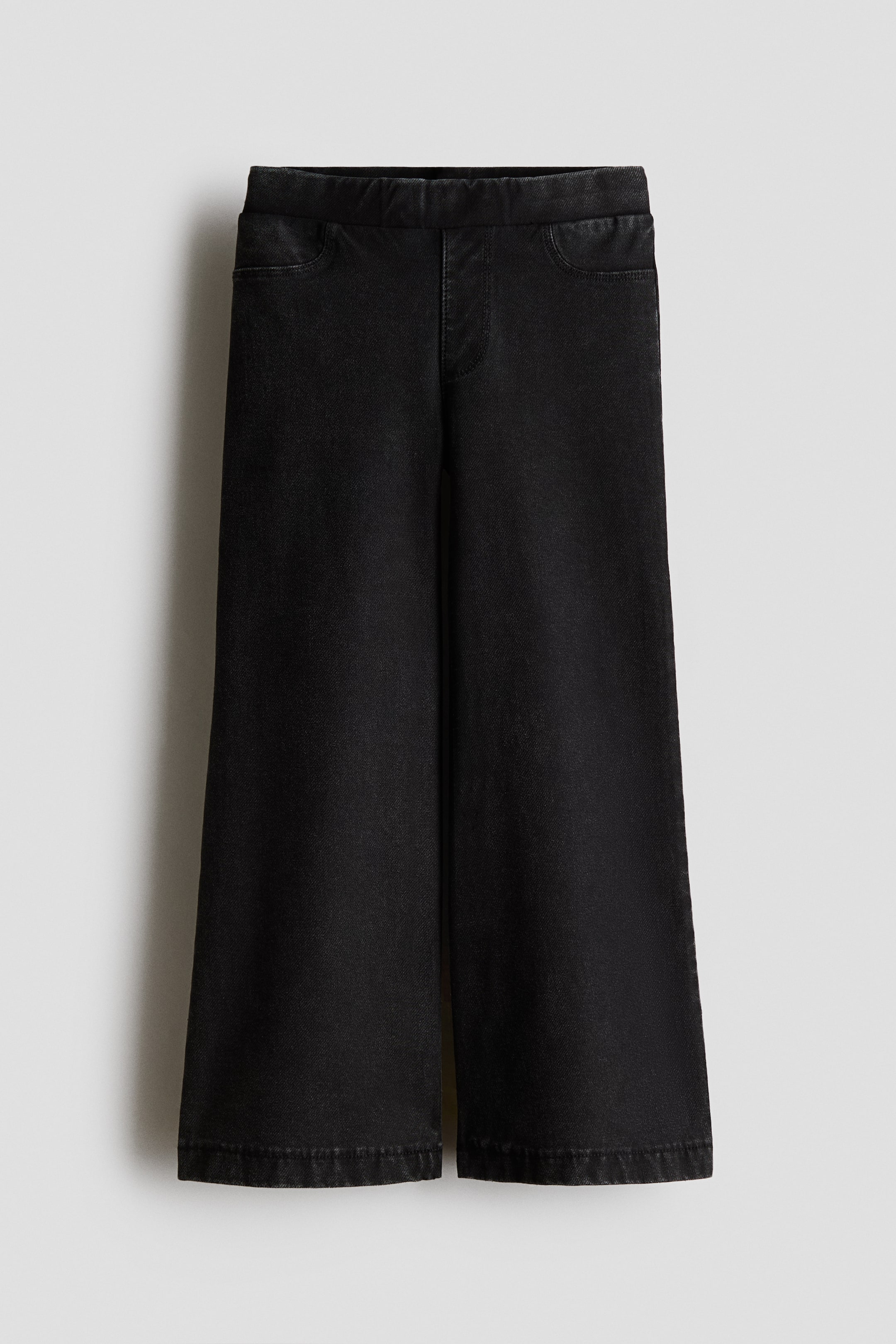 View larger image: A pair of black denim-look jersey jeggings is hanging in a front view, showcasing a wide-leg silhouette. The jeggings feature a flat elasticized waistband, a mock fly, and decorative curved front pockets.