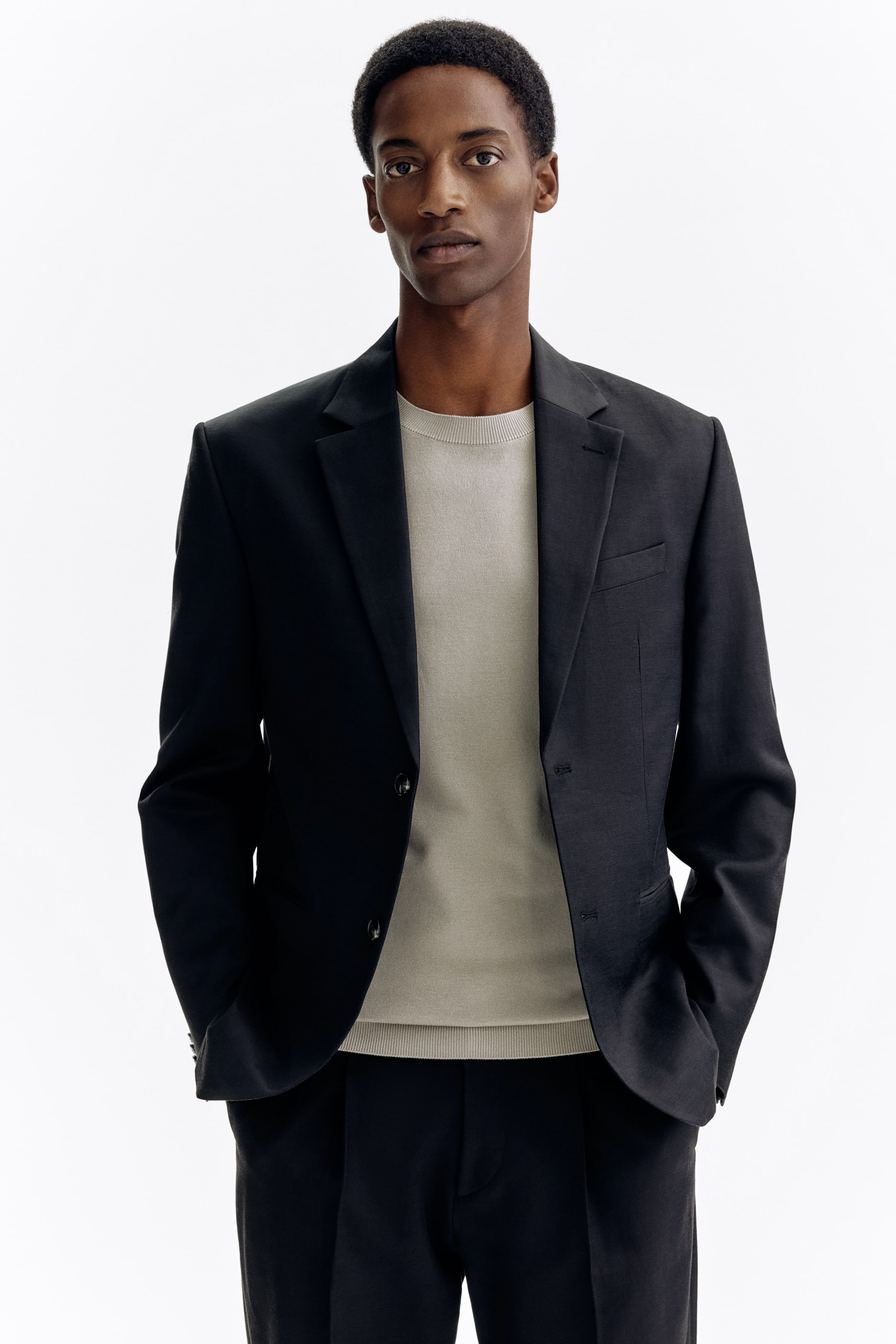 View larger image: The black blazer showcases notch lapels, a chest pocket, a two-button front closure, and flap pockets. It is styled open over a light-coloured crew-neck sweater and dark trousers.