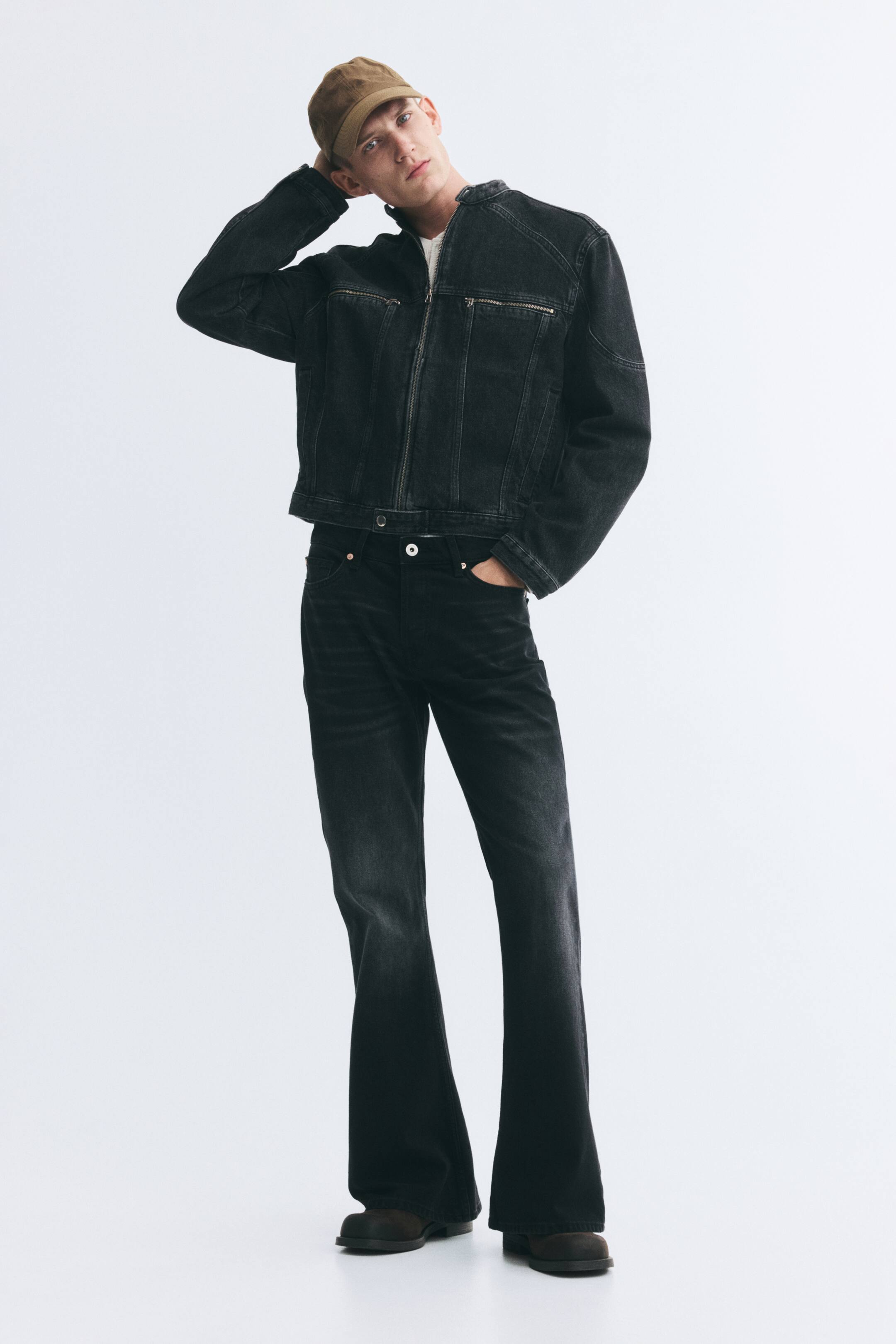 Ver imagen más grande: La modelo lleva una chaqueta corta de denim negro rígido de algodón, con cuello mao, cierre frontal de cremallera y bolsillos en el pecho con cremallera. La chaqueta se combina con una camiseta blanca y unos vaqueros negros a juego, mientras la modelo posa con una mano apoyada sobre una gorra.