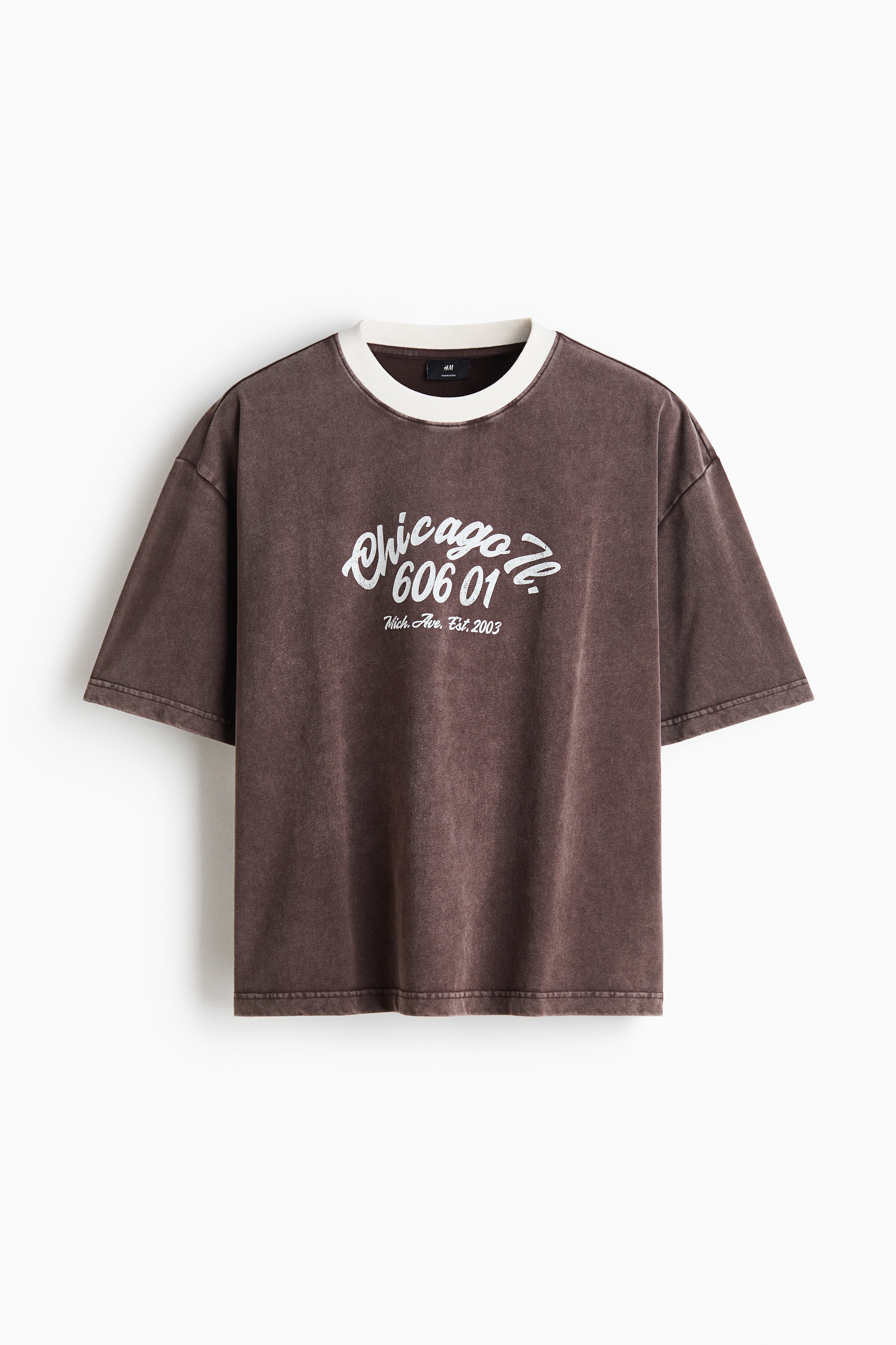 モデルは、ヘビーウェイトのコットンジャージー素材を使用した、色褪せたブラウンのオーバーサイズTシャツを着用しています。フロントには白のグラフィックプリントをあしらい、ドロップショルダーとコントラストの効いた白いリブネックが特徴です。ダークジーンズと色褪せたレッドのキャップを合わせてスタイリングしています。