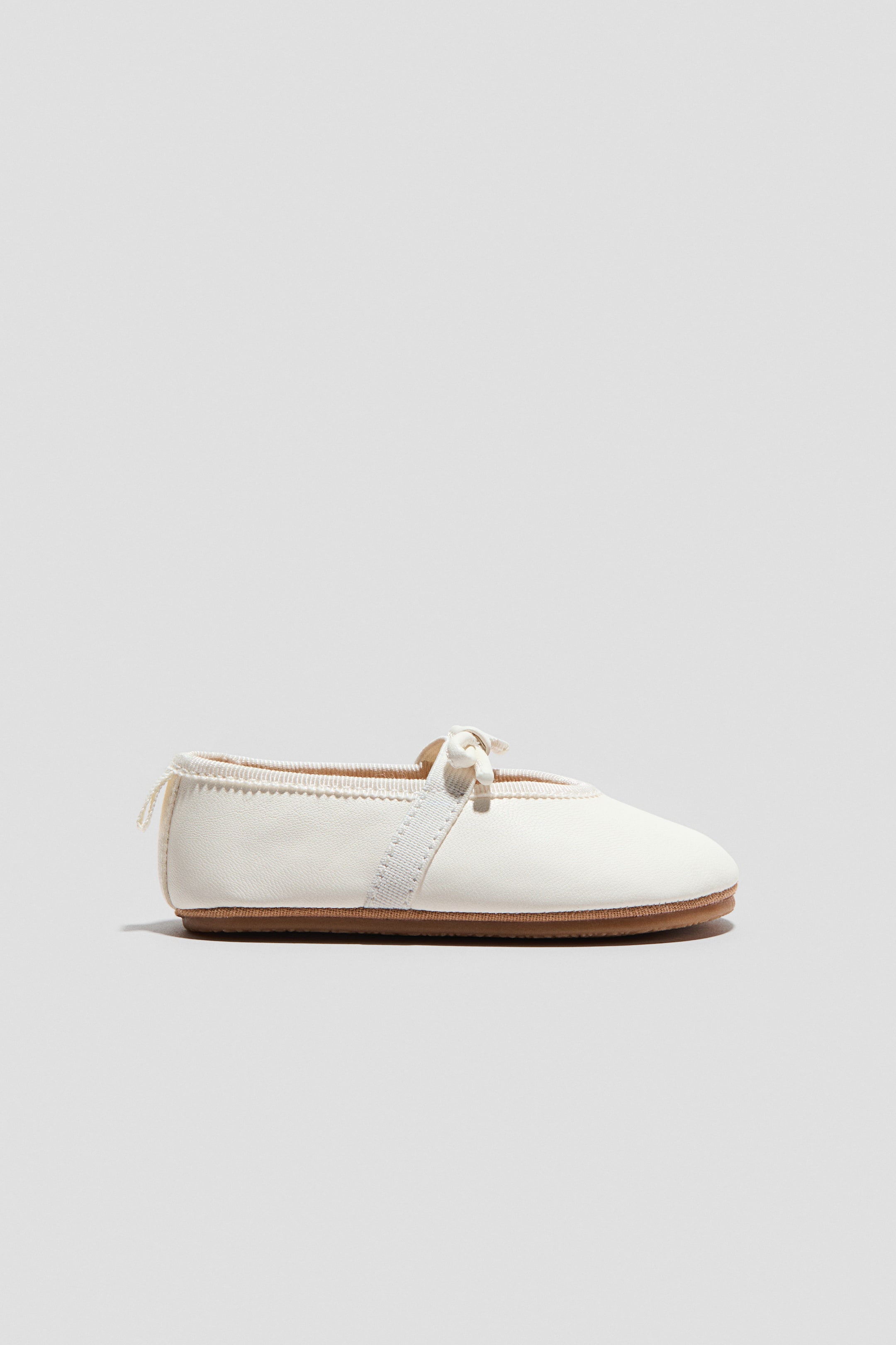 View larger image: An off-white ballet pump is showcasing its side profile, highlighting a rounded toe. A grosgrain strap elegantly crosses over the foot, tying into a neat bow, while a grosgrain loop is visible at the back, resting on a flat brown sole.