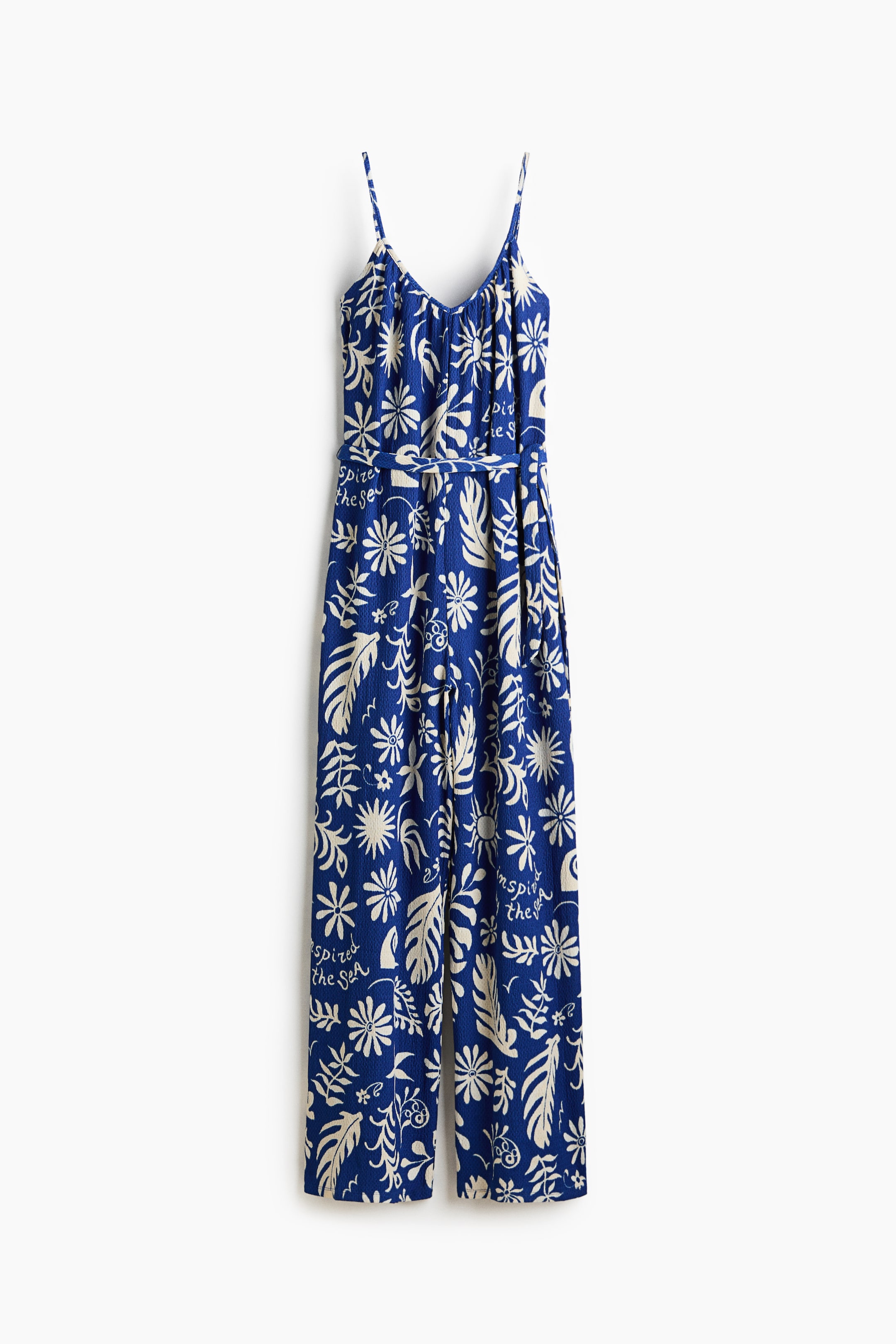 Grotere afbeelding bekijken: De jumpsuit is uitgevoerd in een blauwe gekreukte jersey met een all-over crème bloemen- en bladdessin, inclusief de tekst 'inspire the sea', zichtbaar vanaf de voorkant. Het ontwerp heeft smalle, verstelbare schouderbandjes, een V-vormige halslijn met plooien aan de voorkant, een brede afneembare tailleband en rechte pijpen.