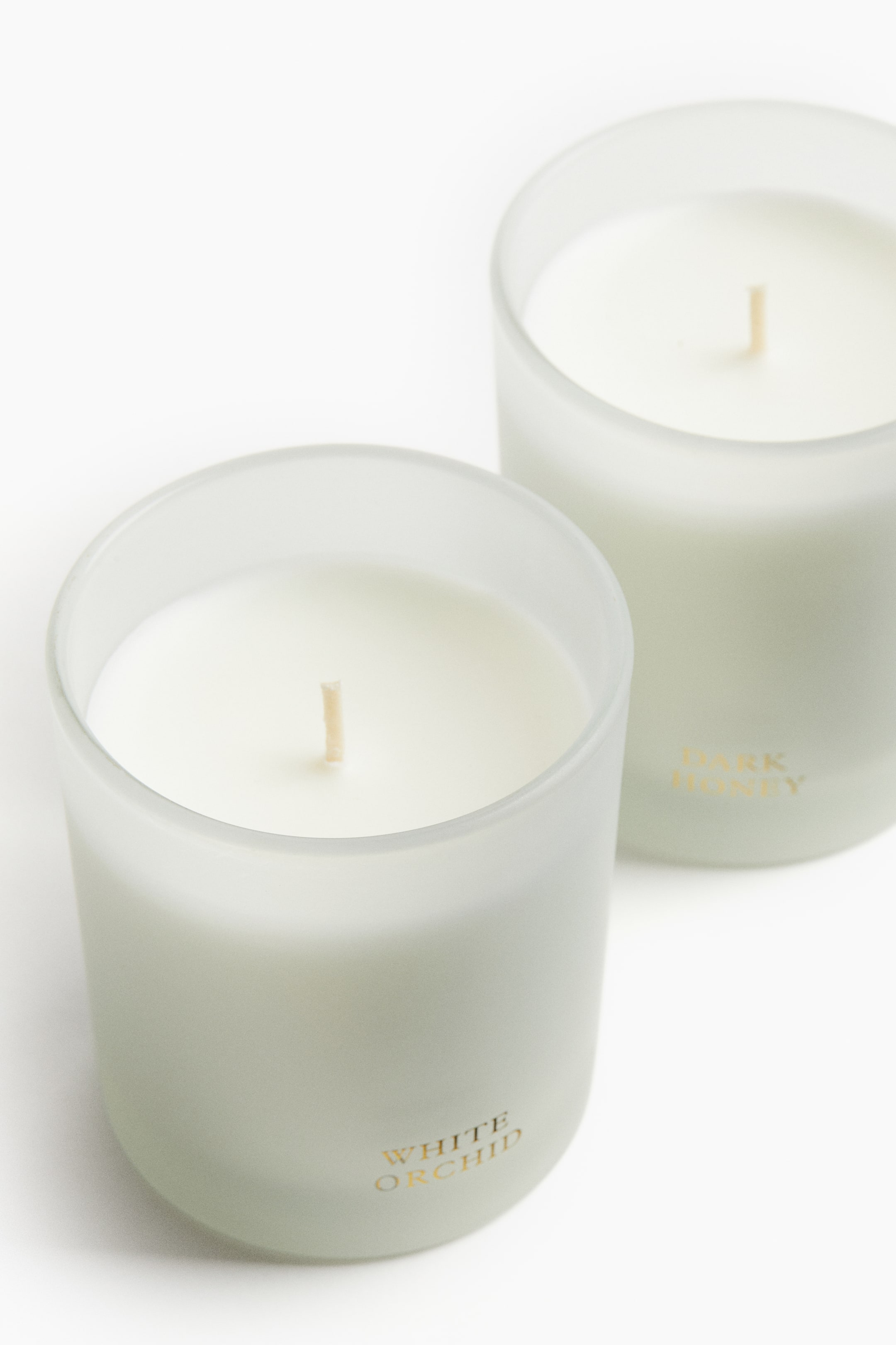 View larger image: Two white candles are sitting in frosted, cylindrical glass holders. Each candle features a central, upright wick, and gold lettering is visible on the front of both holders, reading "WHITE ORCHID" on the front candle and "DARK HONEY" on the one positioned behind it.