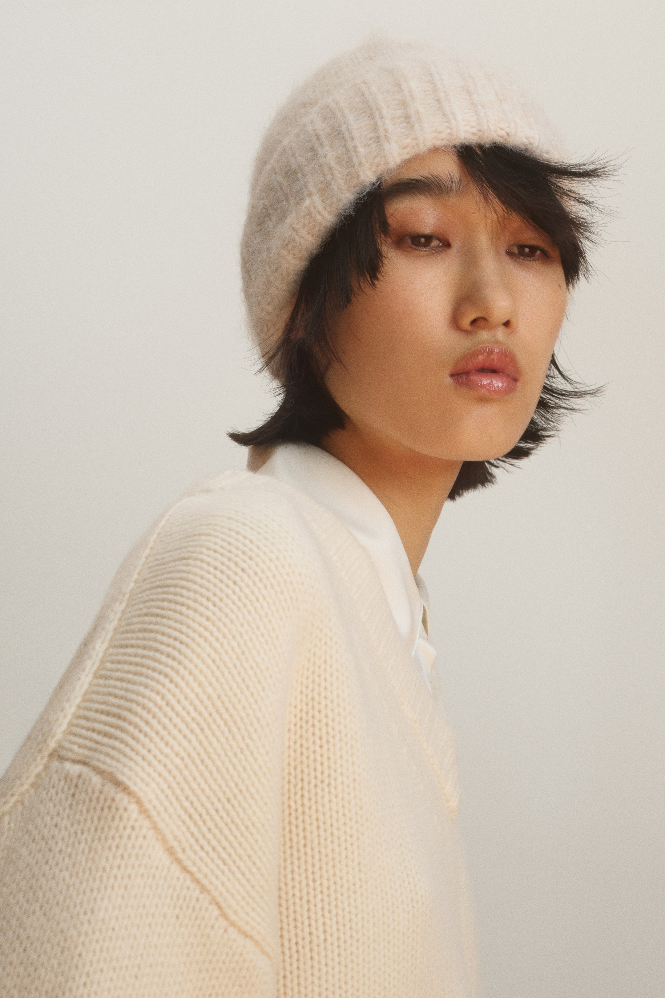 View larger image: A cream-coloured beanie in a soft knit with a brushed finish and a ribbed turn-up hem is being worn by the model. The beanie is styled with a coordinating cream-coloured knit sweater layered over a white collared shirt.