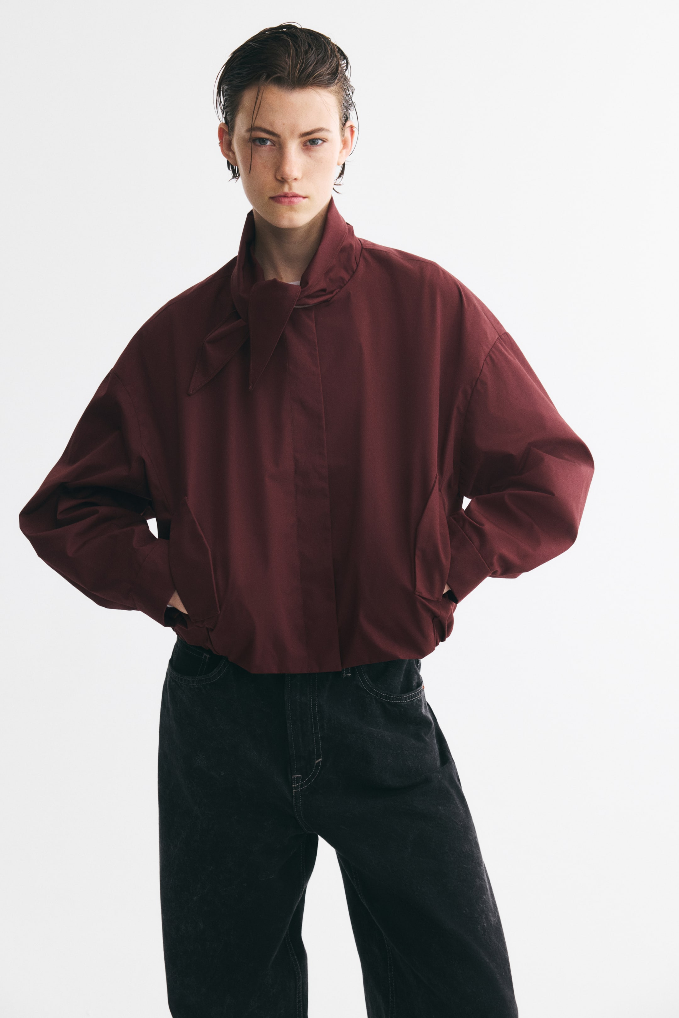 View larger image: A model is wearing a short, oversized burgundy jacket, shown from the front, featuring a high collar with a tied scarf detail at the neck. The jacket is displaying dropped shoulders, long sleeves, and diagonal side pockets with flaps.