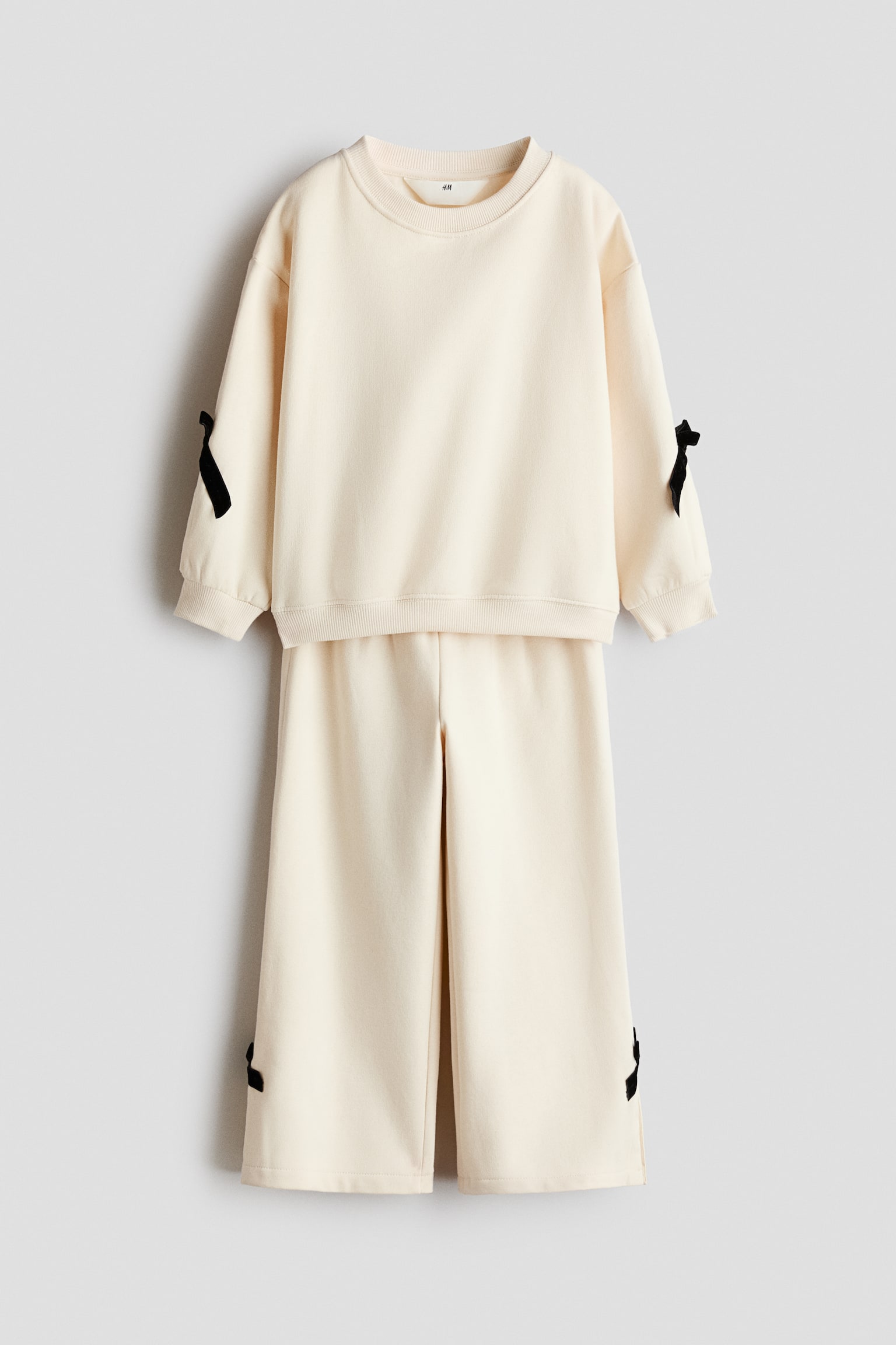 A cream-coloured two-piece loungewear set is shown from the front, consisting of a crew-neck sweatshirt and matching wide-leg pants. The sweatshirt features dropped shoulders with small black decorative bows on the sleeves, while the pants display matching black bows on the lower side hems.