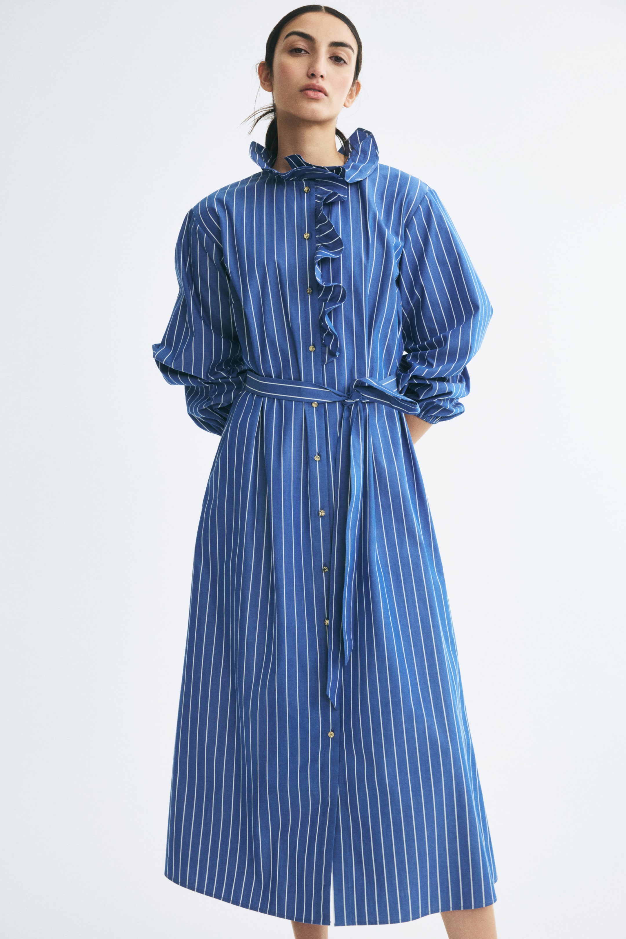 View larger image: The model is wearing a blue and white vertically striped long dress featuring a ruffled collar and matching ruffles along the front button-down placket. The dress is styled with voluminous balloon sleeves and a matching fabric tie belt at the waist.