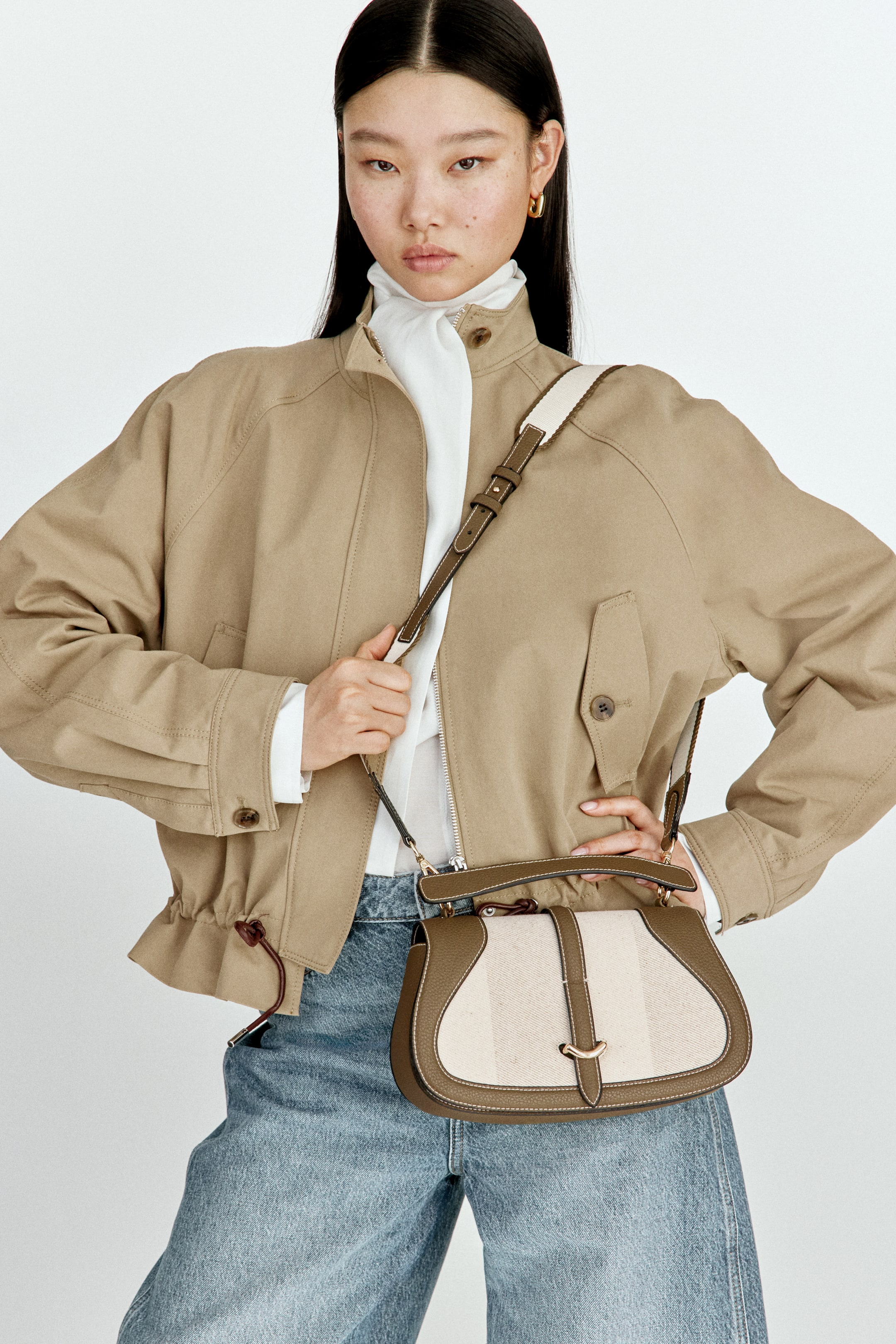 View larger image: The shoulder bag showcases its front with a rounded, saddle-like design, blending a dark khaki-brown leather-look trim with a light-hued, subtly striped fabric body. A gold-toned clasp secures the front flap, complemented by a matching adjustable strap and a small top handle.