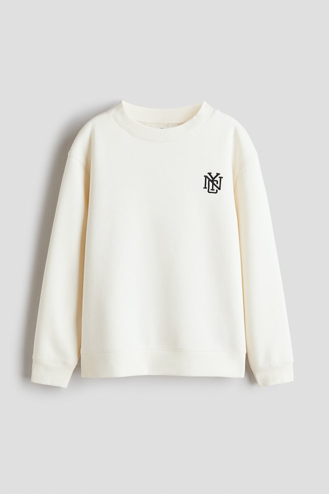 A cream-coloured sweatshirt is shown from the front, featuring a small black motif of intertwined letters on the upper right chest. It is designed with a round, rib-trimmed neckline, dropped shoulders, long sleeves, and ribbed cuffs and hem. - 1