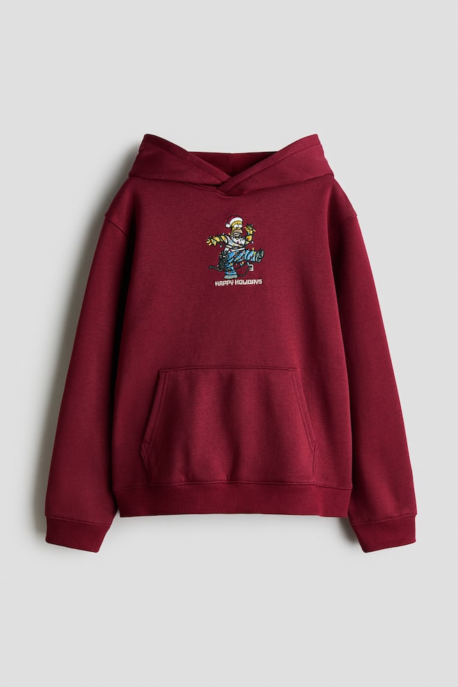 En framifrÃ¥nvy av en vinrÃ¶d fleecehoodie visar en omlotthuva, sÃ¤nkta axlar och en kÃ¤nguruficka. PÃ¥ brÃ¶stet syns ett tryckt motiv av en seriefigur som trasslat in sig i julbelysning ovanfÃ¶r texten "HAPPY HOLIDAYS", med ribbade detaljer vid Ã¤rmslut och nederkant. - 1