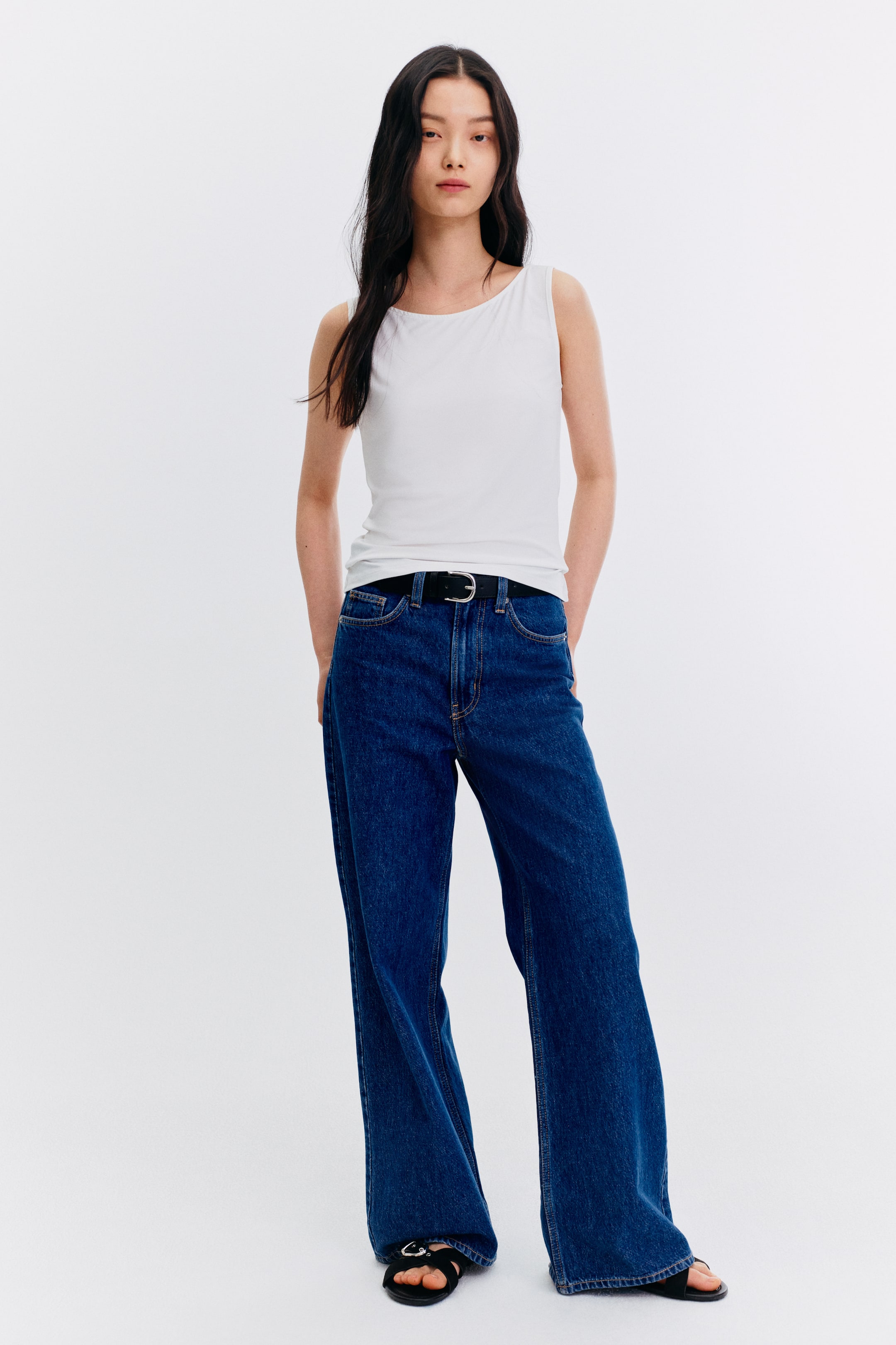 View larger image: The model is wearing a white sleeveless top featuring a wide, rounded neckline. This top fits snugly against the body and is paired with dark wash wide-leg jeans and a black belt.